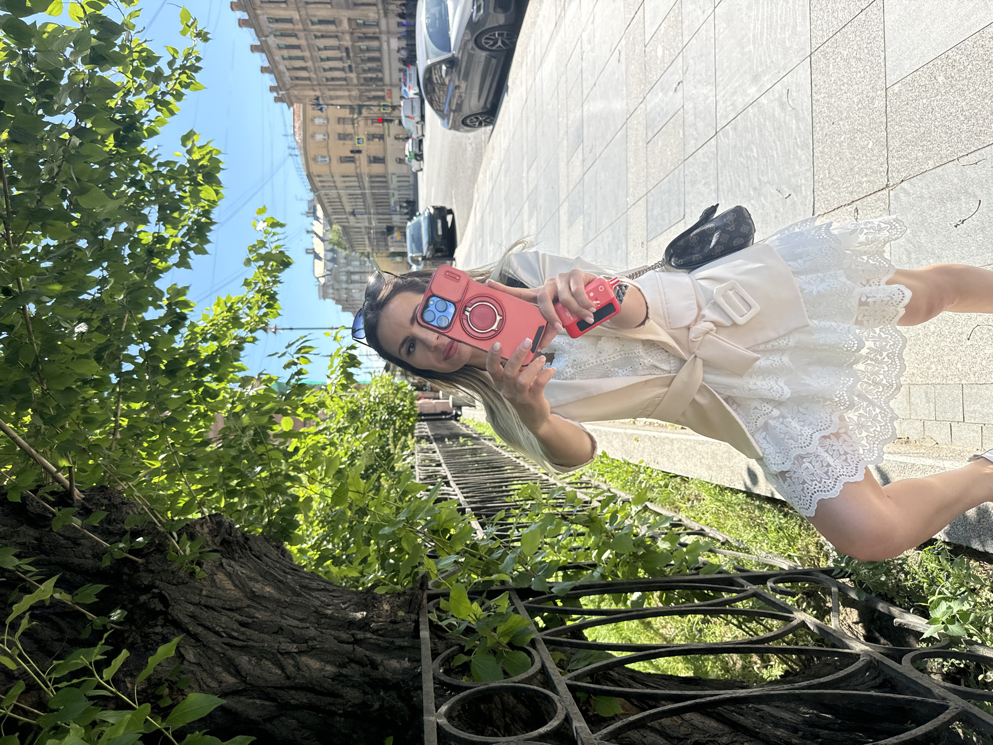 A woman is taking a selfie on a sunny day in Saint Petersburg, Russia. She is standing on a sidewalk next to a tree and an ornate metal fence. She is wearing a light-colored trench coat over a white lace dress and holding a red phone in her left hand, with a power bank attached to it. The phone has a pink case with a ring holder. She is also wearing sunglasses on her head. In the background, there are cars parked along the street and historic buildings with classical architecture. The sky is blue and clear. The scene appears to be a public area, possibly a park or a pedestrian street, given the presence of trees, benches, and the architectural style of the buildings. The woman's expression is neutral. The time of day appears to be midday or early afternoon due to the strong sunlight and shadows.