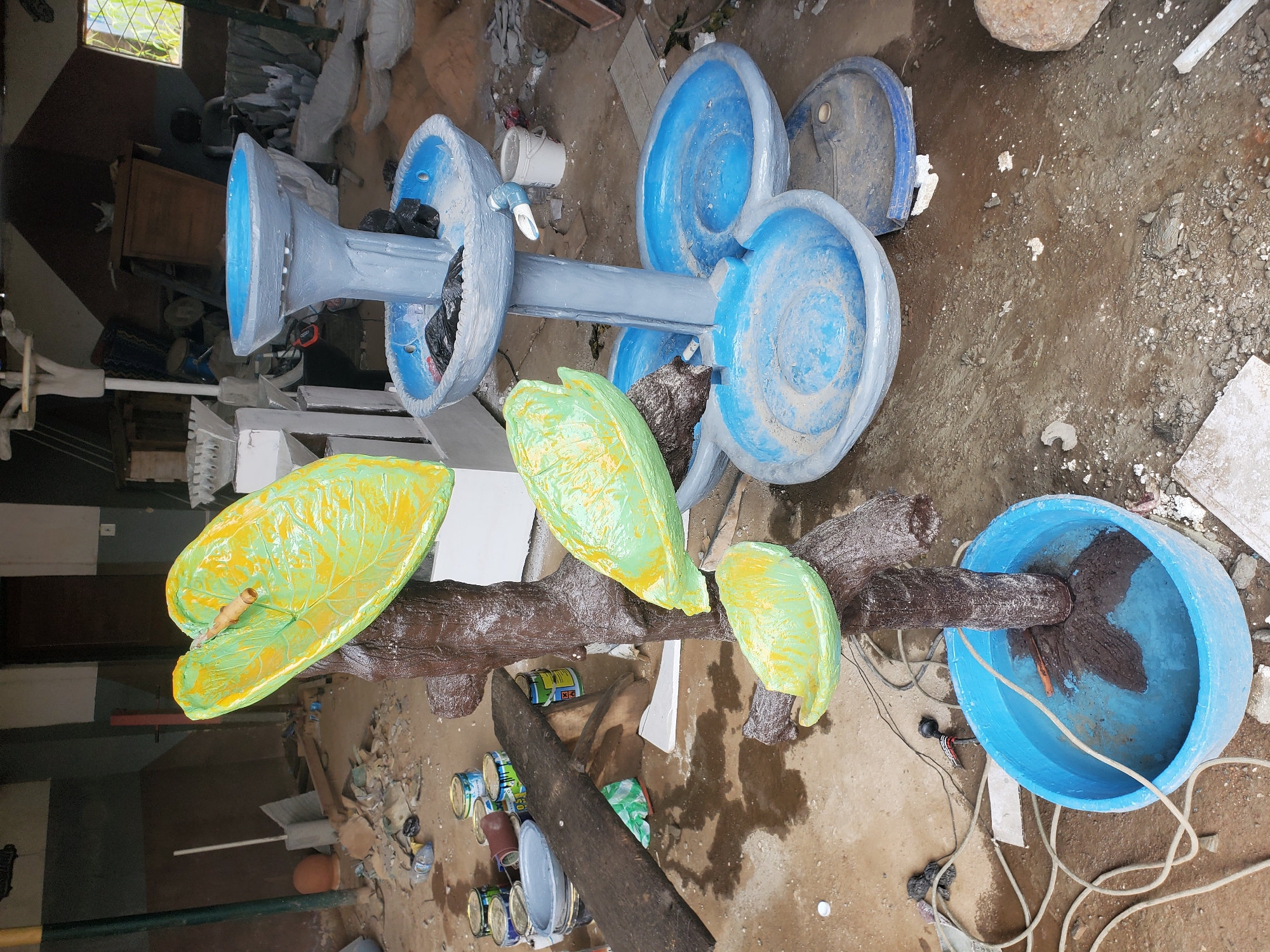 The image shows a partially assembled, multi-tiered fountain structure in a cluttered outdoor environment. The fountain's base is a large blue plastic tub filled with water, with a dark, tree-like support emerging from it. Attached to this support are three decorative, leaf-shaped elements painted in bright green with yellow accents. The fountain's upper tiers are constructed from gray material, with at least two bowls designed to hold water. A white pipe extends from one of the upper tiers.
The surrounding area is a rough, unpaved ground surface with scattered debris, including paint cans, wood, plastic sheeting, and various construction materials. In the background, a building with exposed brickwork and a partially open window is visible. The scene appears to be an outdoor workshop or storage area, possibly located in Jos, Nigeria, given the style of the building and the general appearance of the environment. No people are present in the frame. The lighting suggests it is daytime, possibly overcast.
