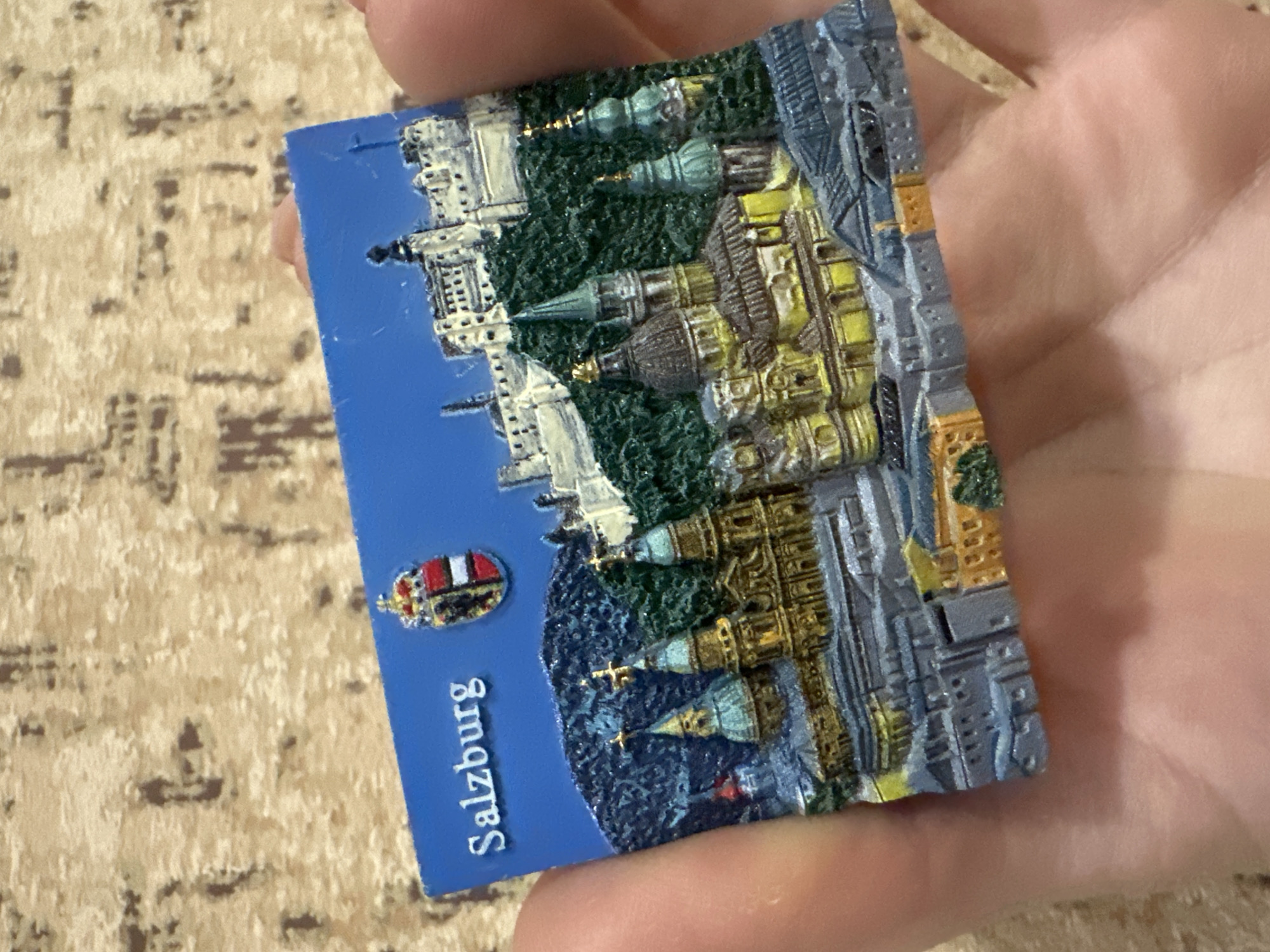 The image shows a souvenir magnet held in a hand. The magnet depicts the city of Salzburg, Austria. The word "Salzburg" is printed on a blue background. The magnet also features miniature depictions of landmarks, including a castle, churches, and other buildings. The details are three-dimensional and painted with various colors. The background is blurred, showing a patterned surface.