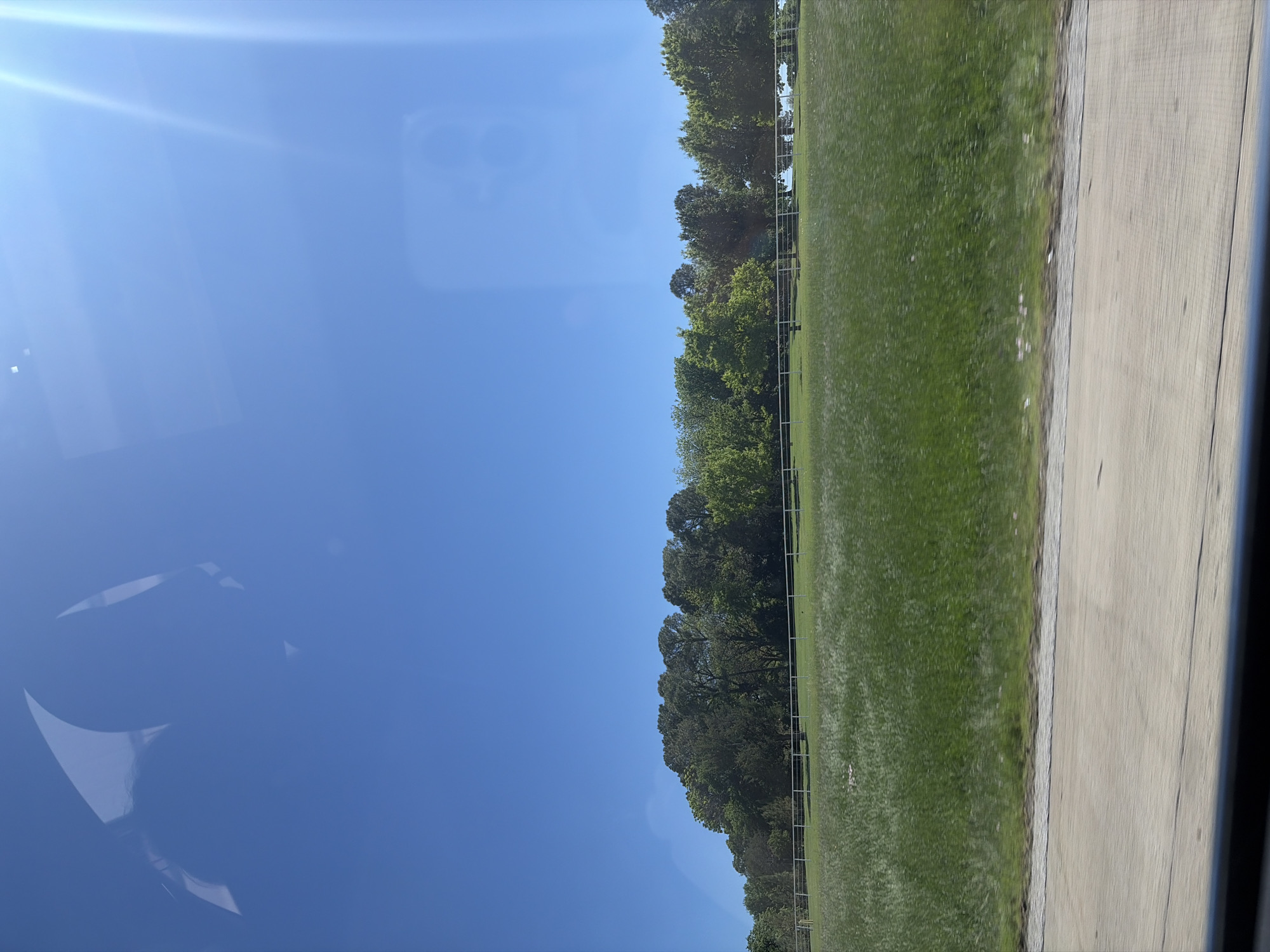 The image presents a landscape as viewed from a moving vehicle. The sky is clear and blue with lens flare, occupying the left side of the frame, with a possible reflection visible on the window. A line of trees separates the sky from a grassy field on the right, which has a fence in front. Next to the field is a concrete road. The location, given the context, is assumed to be somewhere in Westlake, United States of America. There are no people visible in the image.
