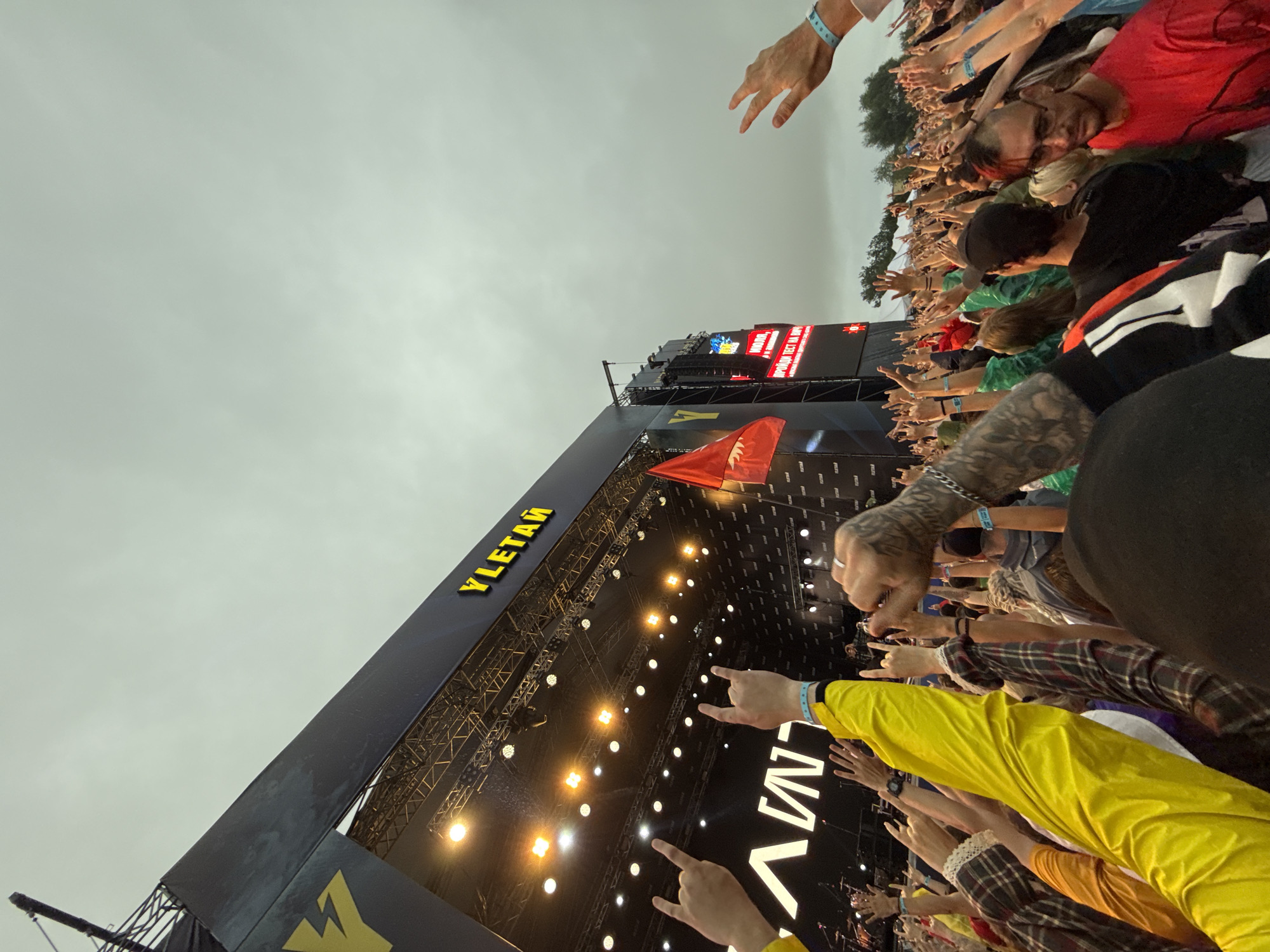 The image depicts a large outdoor concert crowd. The viewpoint is from within the crowd, looking up towards a brightly lit stage. A large banner with yellow lettering reading "YLETАЙ" is visible above the stage. Several people in the foreground are raising their hands, with some making the "rock on" hand gesture. The sky is overcast and appears to be grey. The overall atmosphere suggests a vibrant and energetic event.