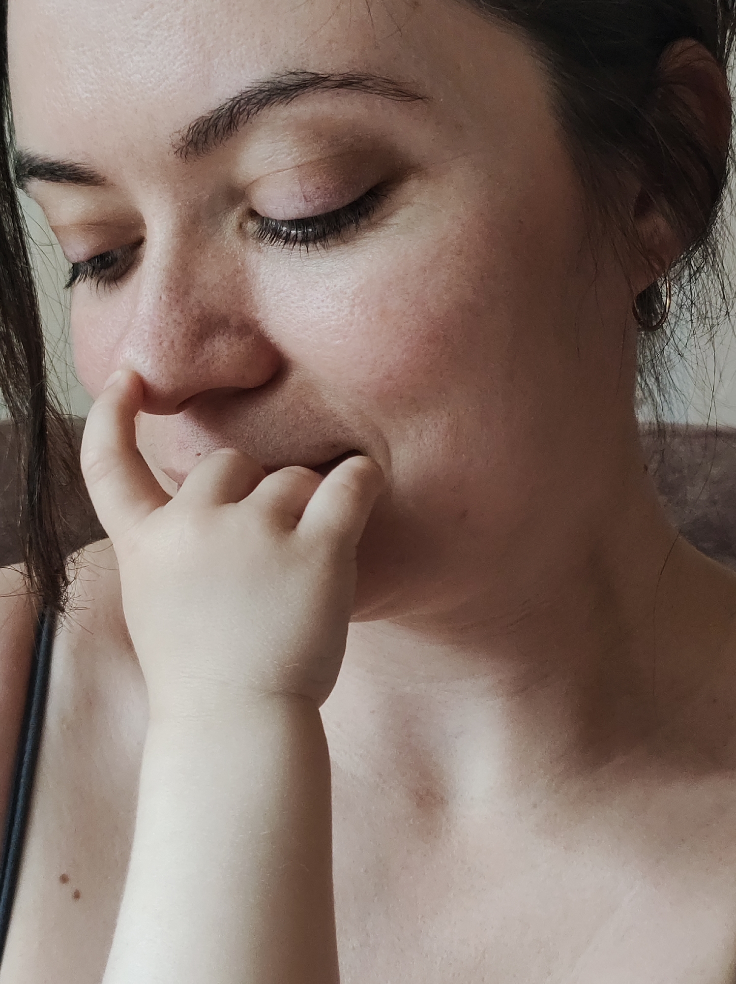 A close-up photograph depicts a woman with fair skin, freckles, and dark hair, likely indoors. Her eyes are closed and looking down. A child's arm and hand are visible in the foreground; the child's finger is touching the woman's nose. The woman's lips are slightly parted, and a gold hoop earring is visible on her left ear. No information suggests this photograph was taken in Chaikovsky, Russia.