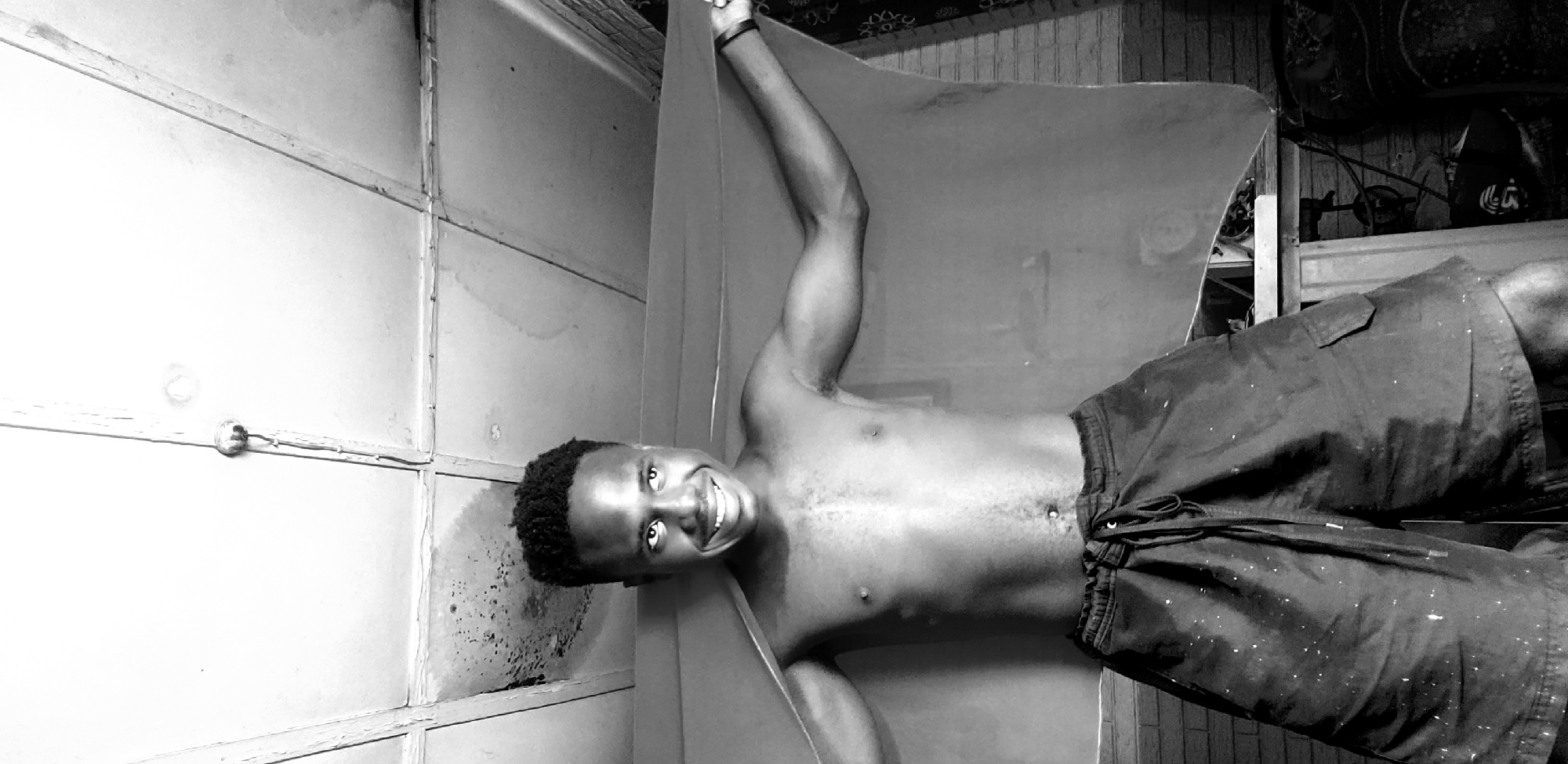 In the city of Narayi, Nigeria, a young Black male is pictured standing shirtless, wearing dark shorts with paint splatters. He is holding up a light-colored cloth with his arms extended to the sides, creating a backdrop. The man is smiling and looking to his right. The environment appears to be indoors, with a textured ceiling overhead that has visible lines and some water staining. A light fixture is present on the ceiling. In the background, a brick wall is visible, along with some furniture or objects. The image is rendered in black and white, highlighting the textures and contrast within the scene.