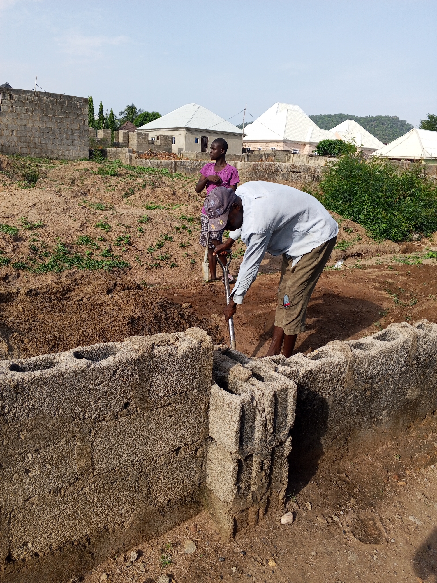 Location: Zango-ndaji, Nigeria.
Scene: Two individuals are present on a construction site. One person is actively digging soil within a rectangular foundation demarcated by stacked concrete blocks. This person is wearing a light-colored long-sleeved shirt, khaki shorts, and a baseball cap. They are using a shovel. The second person, standing behind them, wears a purple shirt and patterned skirt and carries a shoulder bag. Partially constructed buildings are visible in the background, along with light green vegetation and rolling hills. The sky is a clear blue. The ground is predominantly dry, reddish-brown soil.