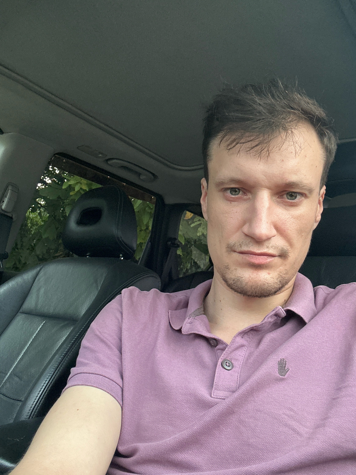 In Shatava, Ukraine, a man is sitting in the driver's seat of a car. The interior of the car is visible, with black leather seats and a sunroof. The man is wearing a purple polo shirt with a small hand logo on the left chest. He has light skin, brown hair, and green eyes. He is looking directly at the camera. Outside the car, greenery is visible through the window, suggesting an outdoor setting, possibly a rural or residential area. The lighting indicates it is daytime.