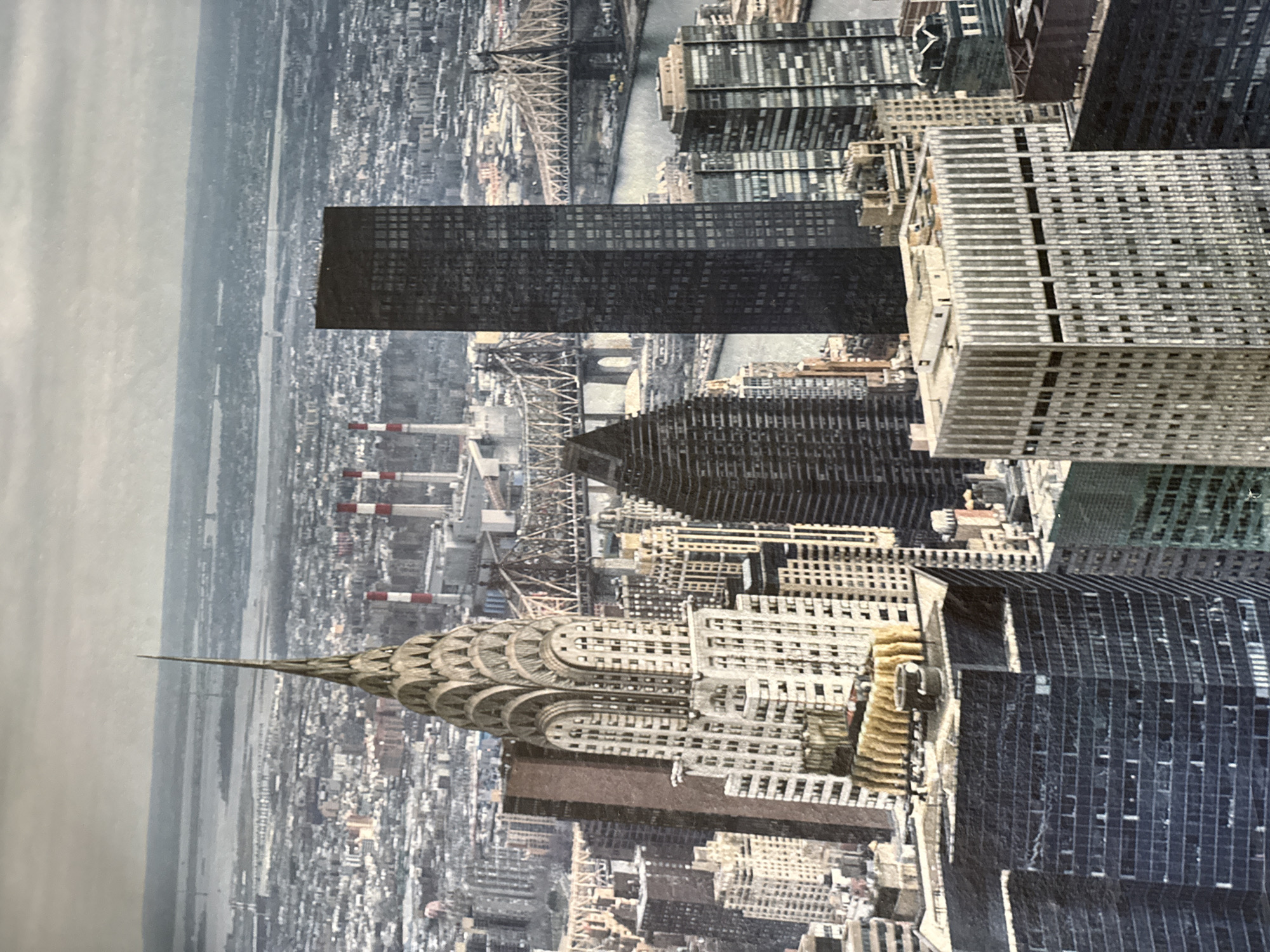 The image depicts a panoramic view of a cityscape, dominated by tall buildings and a body of water. In the foreground, a distinctive art deco skyscraper, the Chrysler Building, stands prominently, its spire piercing the sky. To its right, a dark, rectangular skyscraper rises, its facade covered in windows. Further back, bridges span the water, connecting different parts of the city. The background reveals a sprawling urban landscape with numerous other buildings and what appears to be a power plant with red and white smokestacks. The sky is overcast, casting a diffused light over the scene. The image does not contain any indication of the city of Montana, Bulgaria, and the architectural style suggests an American city, likely New York City given the presence of the Chrysler Building. There are no people visible in the image.