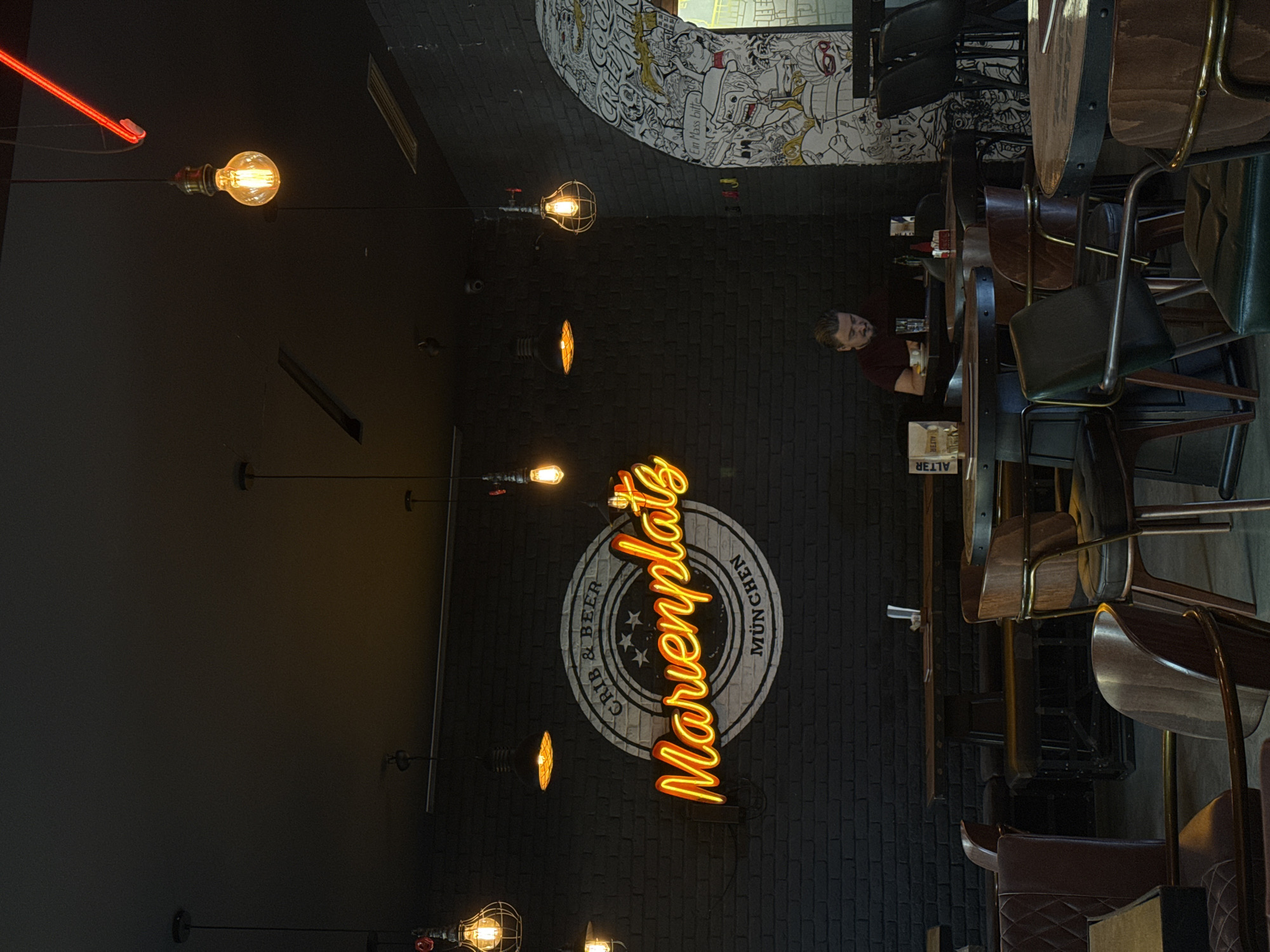 The image captures the interior of a dimly lit bar or restaurant, showcasing a blend of industrial and themed decor. The scene is dominated by dark brick walls and ceiling, from which several exposed incandescent light bulbs, some with wire cages, hang on wires. A bright red neon strip light is visible in the upper left.
A prominent orange neon sign on the left wall vertically spells "Marienplatz." Behind it is a circular logo that reads "CRIP & BEER" at the top and "MÜNCHEN" at the bottom, surrounding three stars, strongly suggesting a German/Bavarian theme for the establishment, despite its location in Podgorica, Montenegro. Further supporting this theme, a black and white cartoon-style mural on a curved wall in the upper right features various figures and the German phrase "Ein Mass bitte."
In the right half of the image, several dark wooden tables with metal bases and matching chairs and bar stools are arranged. A single man with short hair and a beard, wearing a dark red shirt, is seated at one of the tables, looking down, possibly at a drink in a glass or a phone. On his table, a red bottle (likely a condiment) and a small white sign with "ALT3R" printed on it are visible. The overall ambiance, characterized by artificial lighting, suggests it is likely evening or nighttime. The establishment appears clean and ready for patrons.