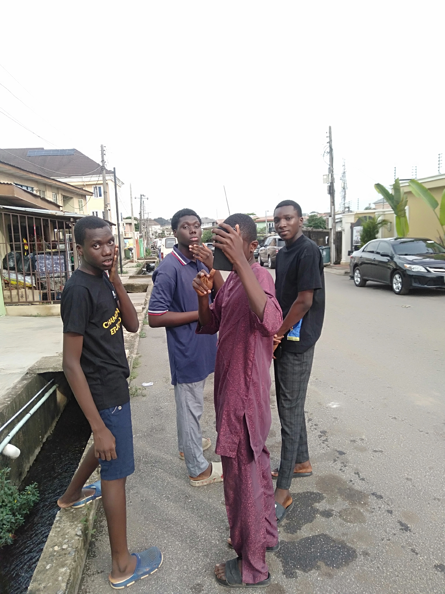 Four young men are standing on a city street in an urban environment. The man on the left is wearing a black t-shirt and blue shorts, with blue sandals. The second man wears a dark blue polo shirt with white and red stripes on the collar and sleeves, gray pants, and light-colored sandals. The third man is holding a tablet in front of his face and wearing a maroon long-sleeved robe with dark sandals. The fourth man wears a black t-shirt, patterned gray pants, and dark sandals. Behind the men, there are residential buildings and parked cars. The street appears to be paved and slightly wet. Overhead power lines are visible.