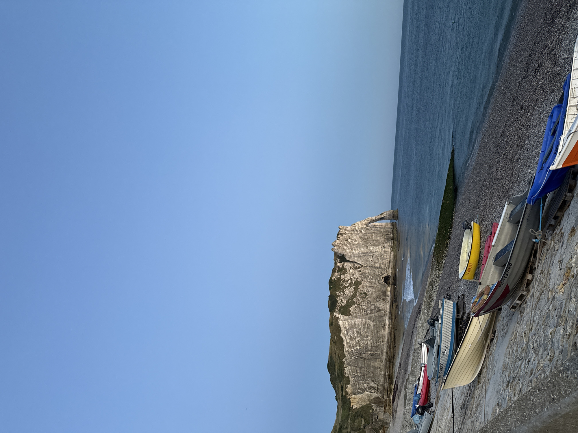 This is a photograph of the pebble beach in Étretat, France, with the famous chalk cliffs and arch of the Falaise d'Aval in the background. Several small boats are pulled up onto the beach. The sky is clear and blue, and the sea is calm. The lighting suggests it is either early morning or late afternoon. There are no people visible in the image.