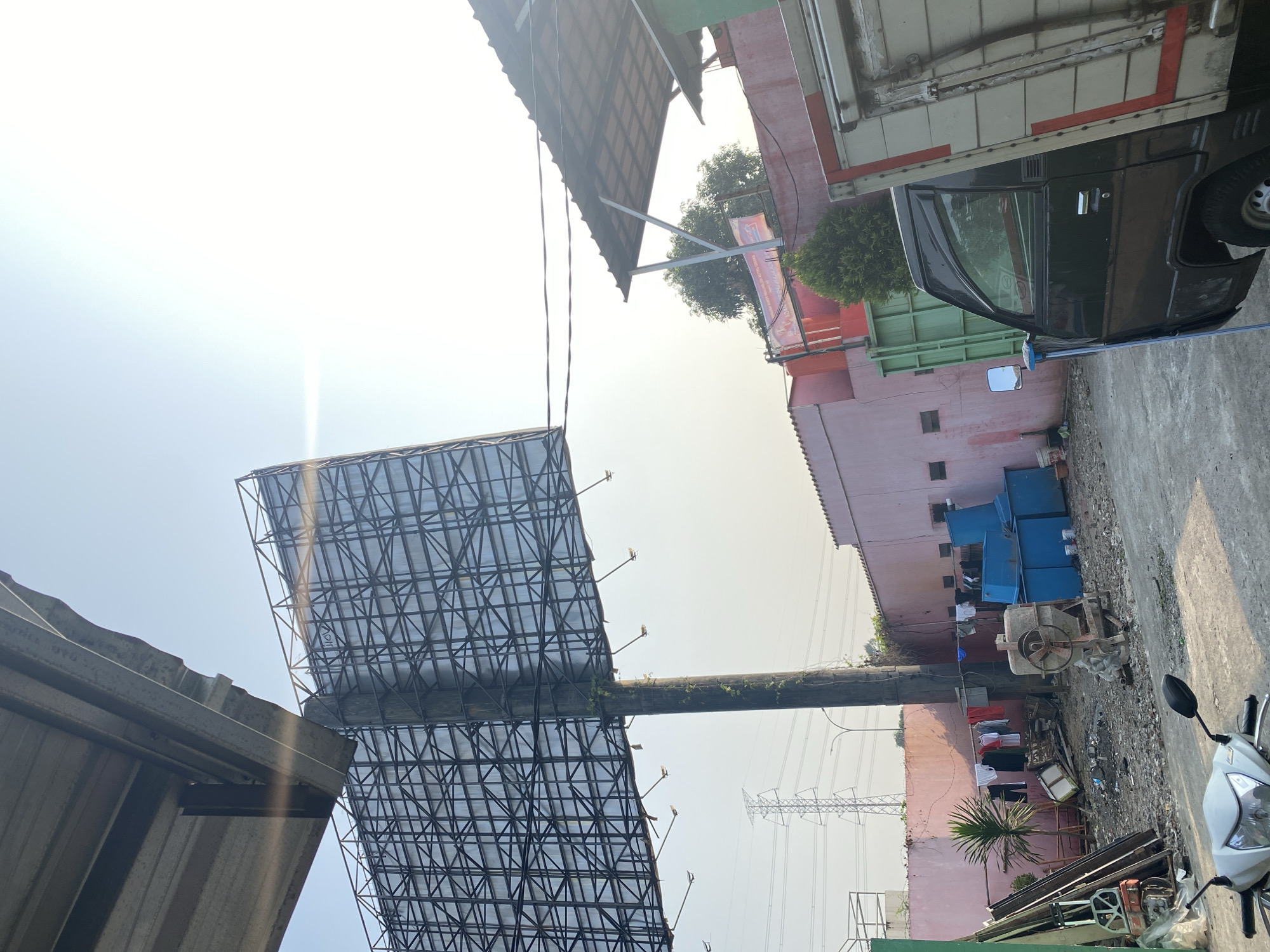 In Tangerang, Indonesia, an outdoor scene unfolds under a bright sky. Dominating the view is the back of a large, dark metal billboard structure supported by a thick, weathered wooden pole. The billboard's frame is a complex grid of metal beams, with a few spotlights attached to its underside. To the left, a portion of a corrugated metal roof juts out, casting a shadow that partially illuminates the billboard's structure.
Below the billboard, a pink wall stretches horizontally, punctuated by several dark, rectangular windows. In front of this wall, a yard appears cluttered with various objects. There's a small palm tree, clothes hanging on a line strung between poles, and a concrete mixer with a metal drum. Scattered debris, including plastic containers, metal scraps, and buckets, covers the ground. To the right of the mixer, several large, bright blue plastic tanks are visible.
On the right side of the frame, a dark green truck with a silver cargo box is parked. The truck has a side mirror and its windows are tinted. A thin, metallic pole leans against the truck. In the foreground, the front of a white motorcycle is visible, with its handlebars and headlights facing the viewer. In the distance, a large electrical pylon with multiple transmission lines stands against the hazy sky. The overall impression is one of an industrial or service area in a developing urban environment, likely a backyard or storage area.