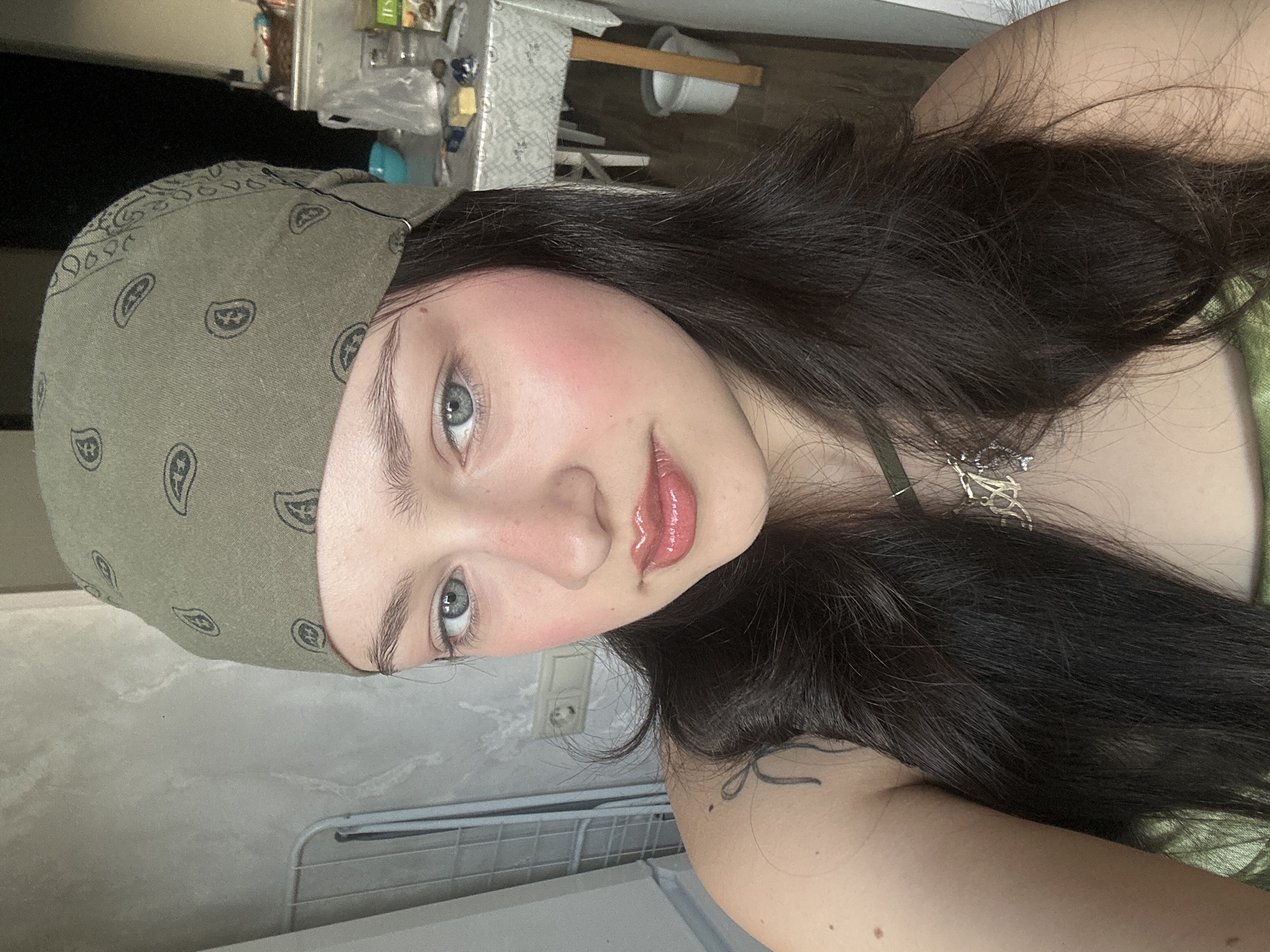 A selfie shows a fair-skinned woman with dark hair and light eyes. She is wearing a green bandana on her head and a shiny lip gloss. She is wearing a necklace. Part of a tattoo is visible on her shoulder. The location is not clear from the image. It is not possible to determine if the photo was taken in Yerevan, Armenia.
