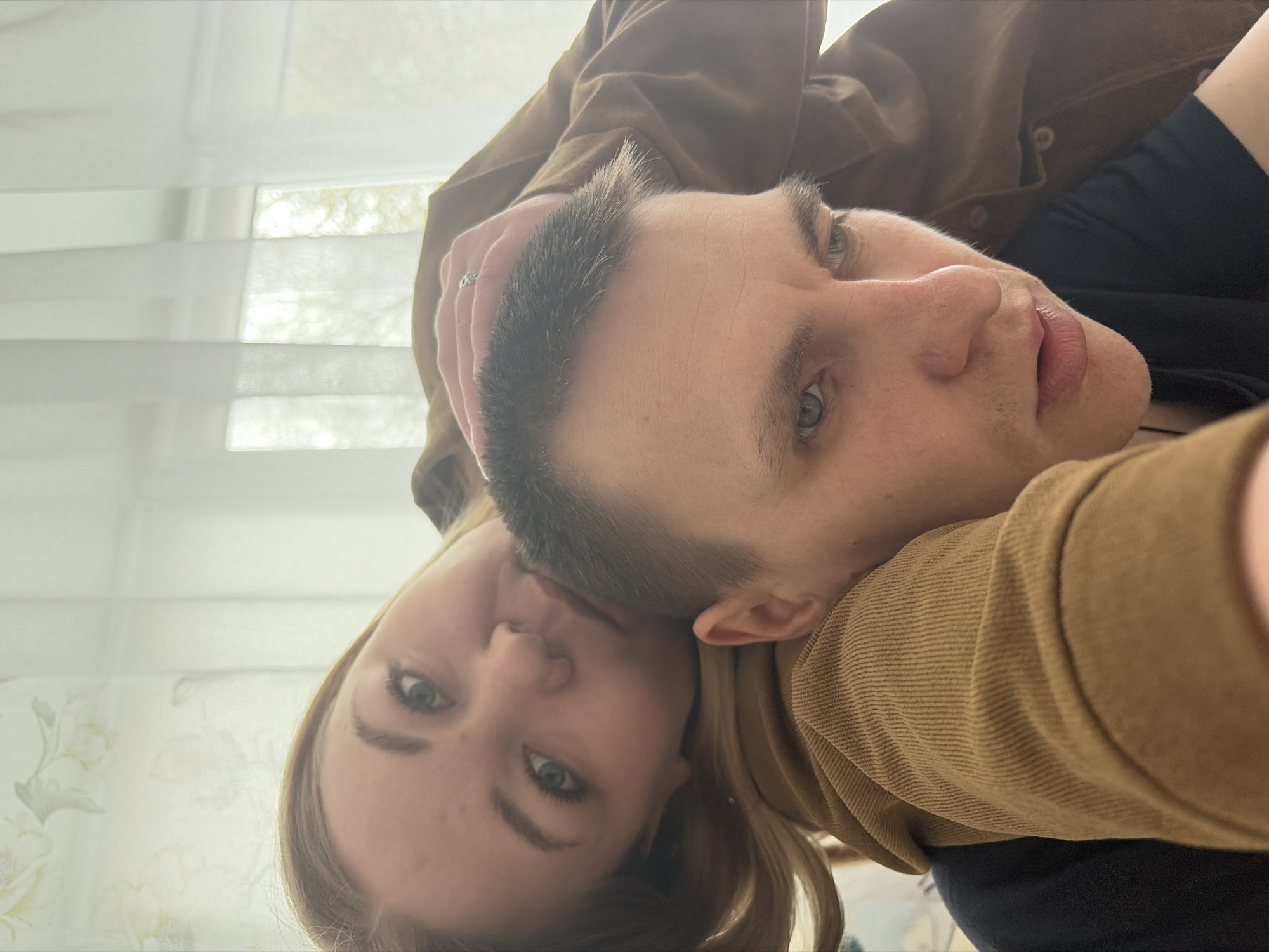 A close-up, rotated selfie features a man and a woman in an intimate pose. The man, with short dark hair, light eyes, and light stubble, dominates the right and central part of the frame. He wears a brown or tan textured (possibly corduroy or ribbed) long-sleeved top over a dark shirt. A silver ring with a decorative band is visible on his finger, held near his head. The woman's face is partially visible to the lower left of the man, with long blonde or light brown hair and light eyes. She appears to be leaning against him, her head resting close to his. Both subjects look towards the camera with neutral to contemplative expressions.
The background reveals an indoor setting, likely a room bathed in bright, diffused daylight, suggesting it's daytime. A window is visible, covered by white horizontal blinds or sheer curtains featuring a subtle floral pattern. The camera angle is from above, looking down on the pair, creating a personal and informal snapshot. The scene conveys a sense of closeness and quiet intimacy. This image is set in Chaikovsky, Russia. There is no visible text in the image.