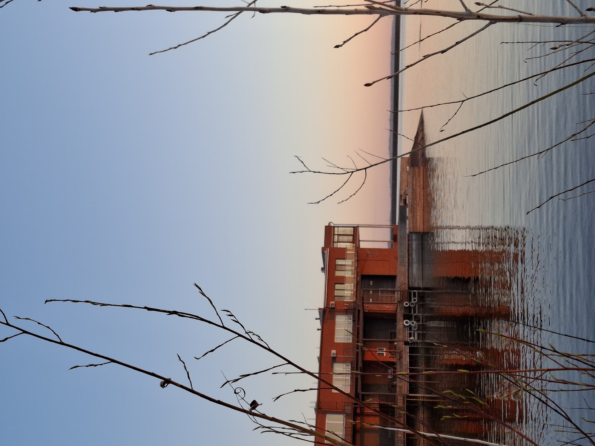 The image depicts a red brick building located in Chaikovsky, Russia, reflected in a body of water. The building has multiple windows and visible metal piping. Branches of a tree frame the scene in the foreground. The sky transitions from blue to orange, suggesting either sunrise or sunset. The water shows distinct ripple patterns, reflecting the building and the sky.