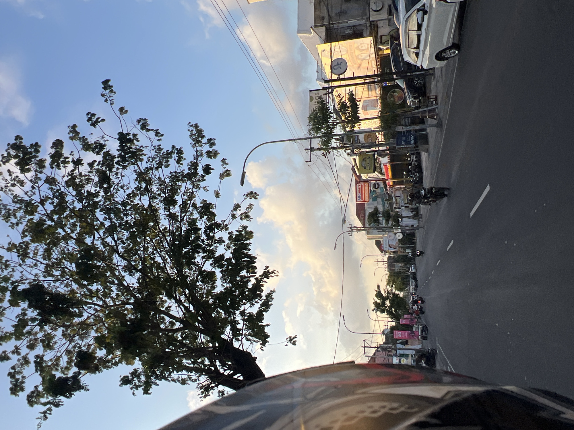 The image shows a street scene in Kuta, Indonesia, likely during the late afternoon or early evening, given the soft light and warm tones in the sky. The perspective suggests the photo was taken from the back of a motorcycle, with the rider's helmet partially visible on the left side of the frame.
The road is paved and appears to be a main thoroughfare with multiple lanes. Several motorcycles and a few cars are visible on the road, indicating moderate traffic. Some of the motorcycles are carrying passengers.
Along both sides of the street are buildings, many of which appear to be shops and commercial establishments. Signs for businesses like "Alfamart" and other commercial entities are visible. There are also streetlights and utility poles with numerous wires.
Trees with green foliage line parts of the street and the sky above is a mix of blue with scattered, soft clouds that are illuminated by the setting sun. The overall atmosphere is that of a busy urban environment.