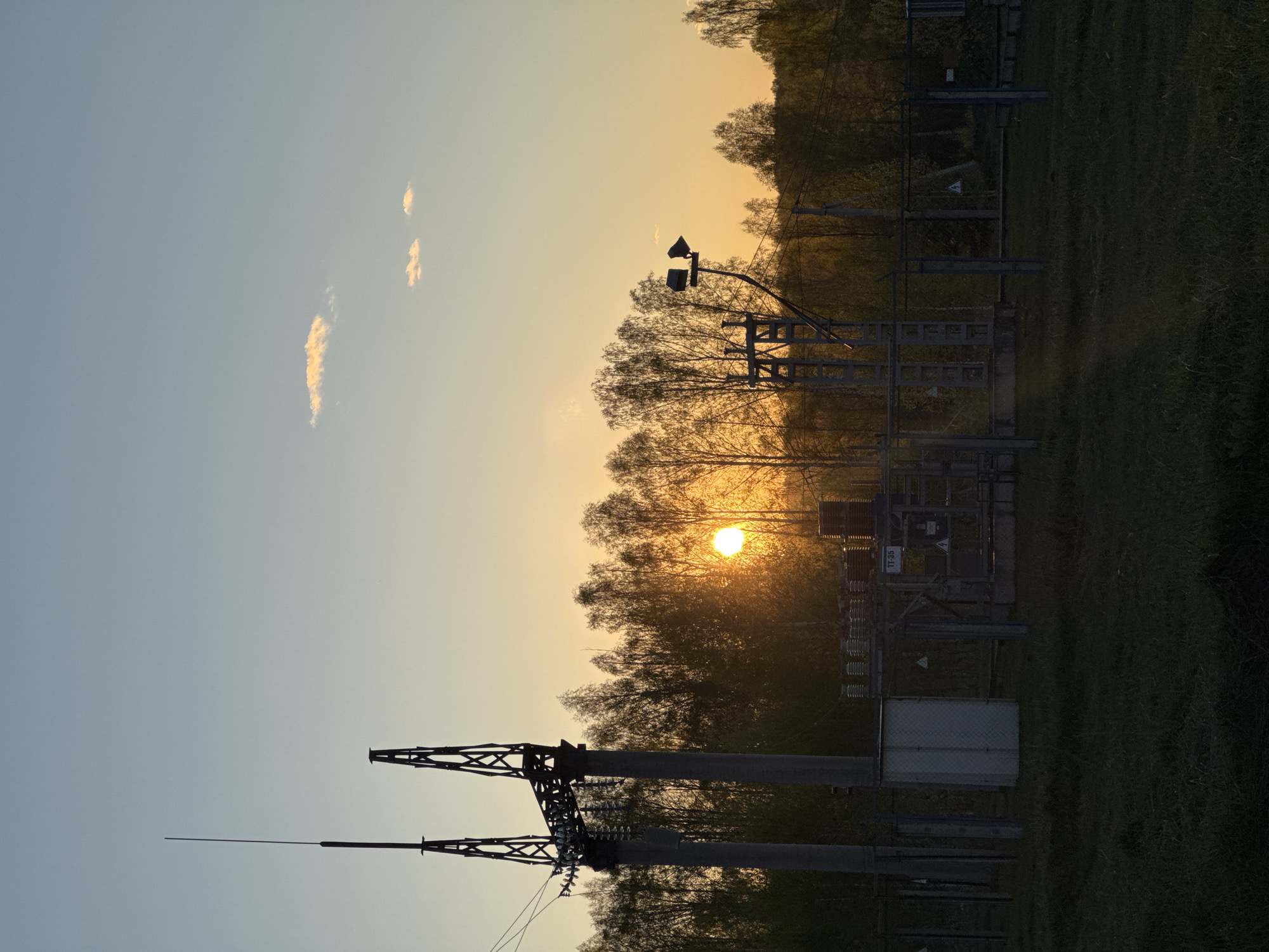 The image shows an electrical substation located in Люкшудья, Russia. The sun is setting behind the substation, creating a silhouette effect. The substation includes metal structures and equipment, including transmission towers and transformers. Trees are present in the background, and there is a grassy field in the foreground. The sky is blue with some clouds.