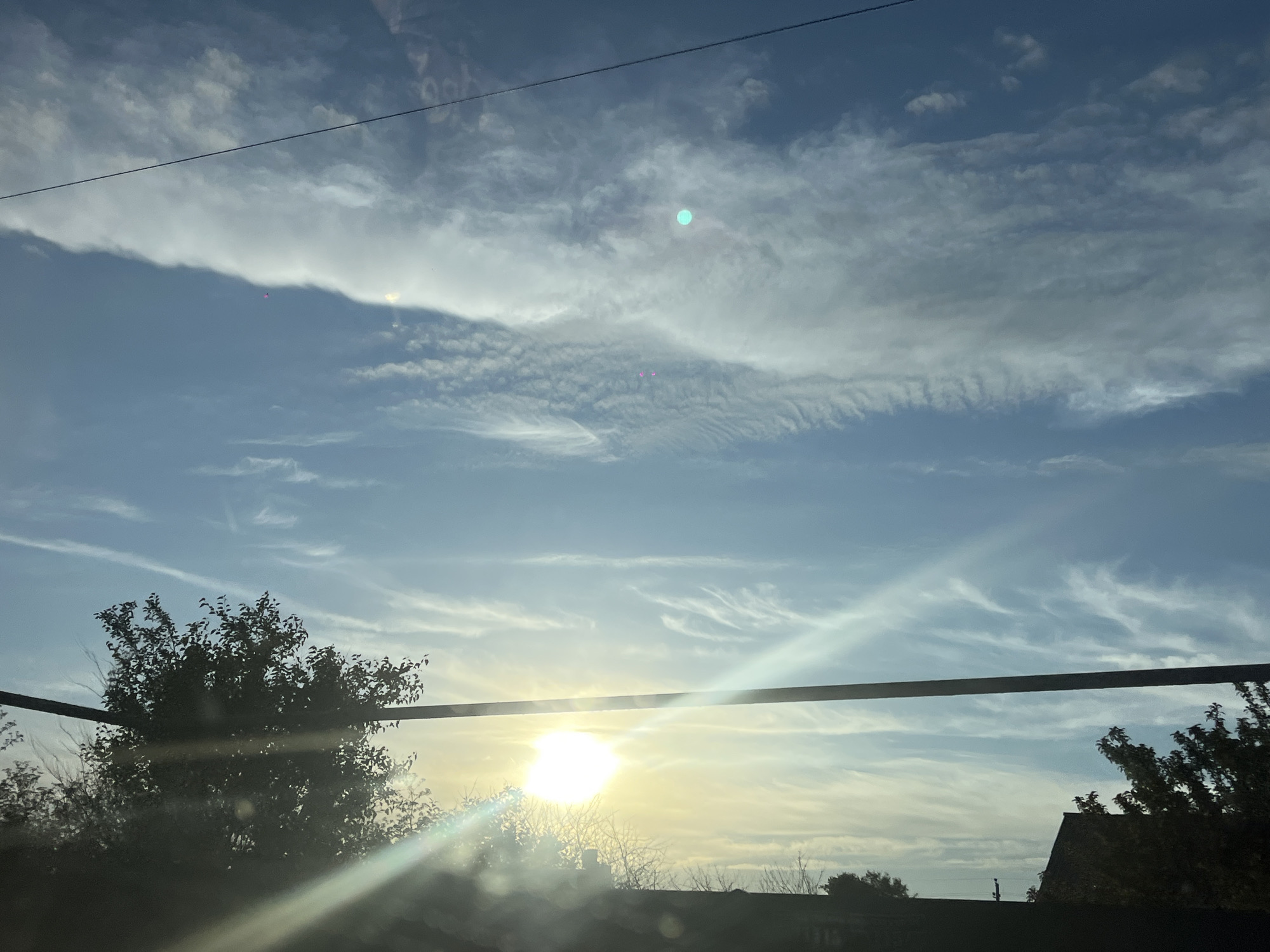 The image captures a daytime sky over Novopokrovskaya, Russia, with the sun prominently visible and casting bright rays. The sky is a blend of blue and white, with various cloud formations, including wispy cirrus clouds and larger, more opaque clouds. A thin black wire stretches horizontally across the upper portion of the image. Below the sky, the silhouettes of trees and rooftops are visible, indicating a semi-urban or suburban environment. The sunlight creates a lens flare effect, particularly in the lower half of the image. There are no people or specific actions depicted.