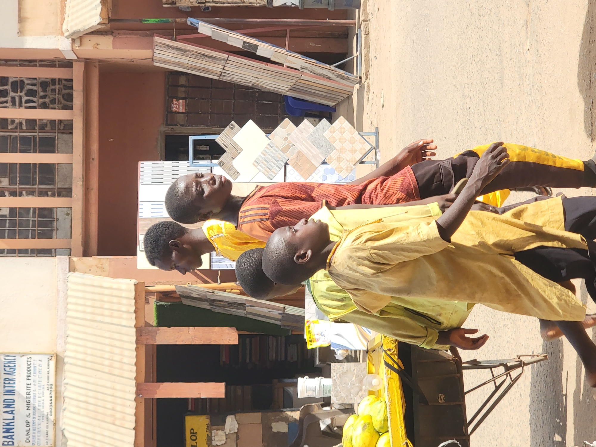 The image captures a scene in Maiburiji, Nigeria, featuring three African men standing along a street. The man closest to the viewer is wearing a yellow shirt and black pants. Behind him is a man wearing a green shirt and behind that man is a man wearing an orange striped shirt and yellow pants. They stand in front of what appears to be a vendor's stall or shop, displaying tiles and other materials, possibly for construction or home improvement. The background reveals buildings, signs, and street items, adding context to the location. A sign reading "BANKLAND INTER AGENCY" can be seen. The environment suggests an urban setting in Maiburiji, with the presence of people, commercial activities, and infrastructure.