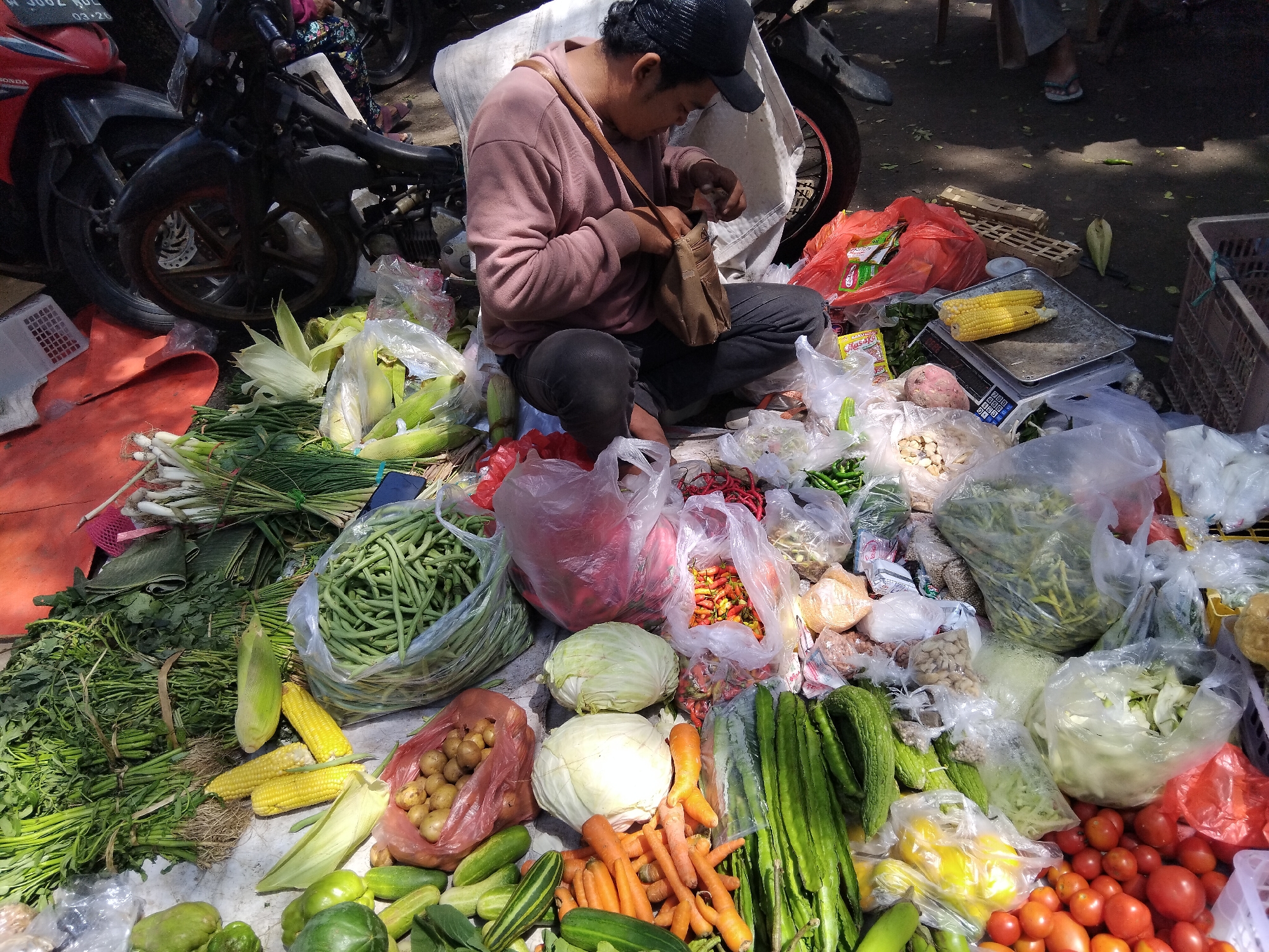 An outdoor market scene in Kepuh Kiriman, Indonesia, shows a person sitting cross-legged among piles of fresh produce. The person, wearing a pink sweatshirt and a black baseball cap, is rummaging through a brown shoulder bag. The ground is covered with a variety of vegetables including long beans, corn on the cob, potatoes, cabbage, carrots, cucumbers, bitter melon, and tomatoes, many of which are packaged in clear plastic bags or red plastic bags. In the background, motorcycles are parked, and there are other market stalls and customers. The lighting suggests a sunny day.