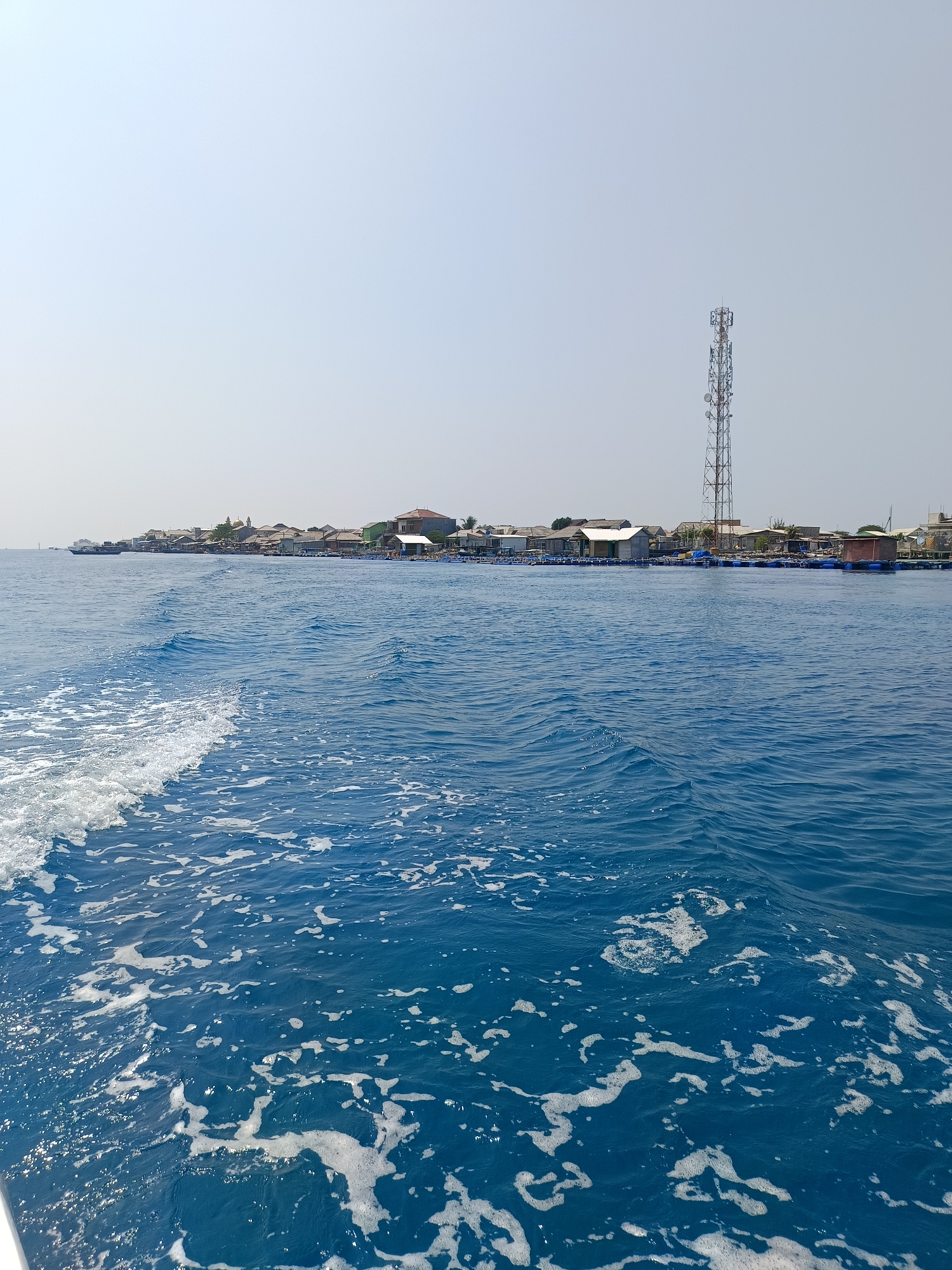 An eye-level, long-shot, outdoor, daytime, low-angle photograph captures the view from the side of a boat moving through blue water. White foam and waves are visible in the foreground, indicating movement. In the distance, a shoreline is lined with numerous buildings, some appearing to be houses and others possibly part of a small settlement. A tall telecommunications tower stands prominently on the shore, and there are structures floating on the water, likely fish farms or aquaculture pens. The sky above is a pale blue, with no clouds visible. The scene suggests a location in or near Jakarta, Indonesia, possibly on the coast or in an archipelago area accessible by boat. No people are clearly visible.