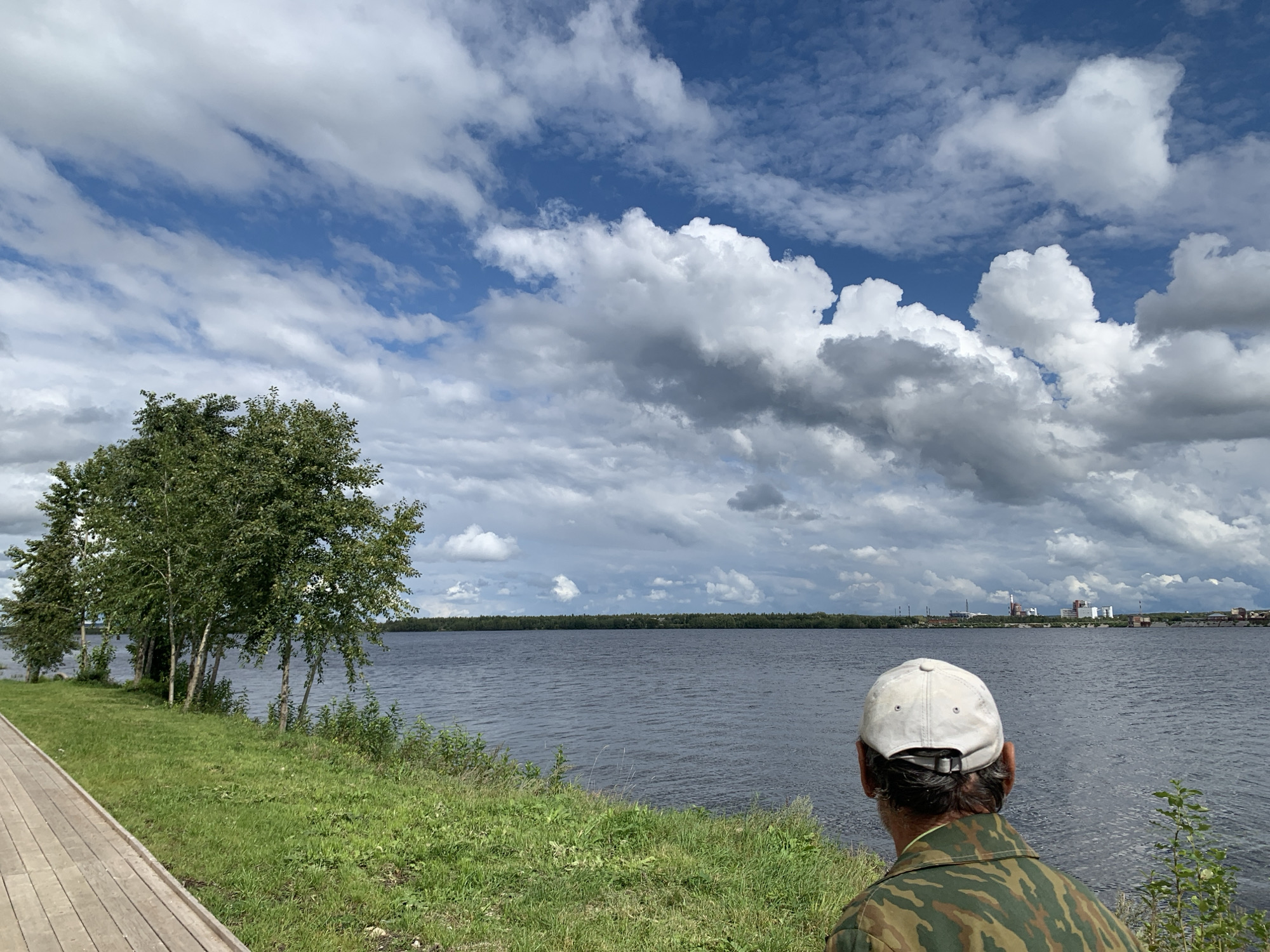 A man wearing a white baseball cap and a camouflage jacket stands facing away from the camera, looking out at a wide, dark body of water. The water appears to be a river or lake, with a distant treeline on the opposite bank. In the far distance, a complex of industrial buildings with tall smokestacks is visible. The sky is a mix of blue and white clouds, with dramatic formations creating shadows on the water. In the foreground, to the left, a wooden boardwalk runs along a grassy bank. The overall scene suggests a man observing an industrial landscape from a waterfront park.