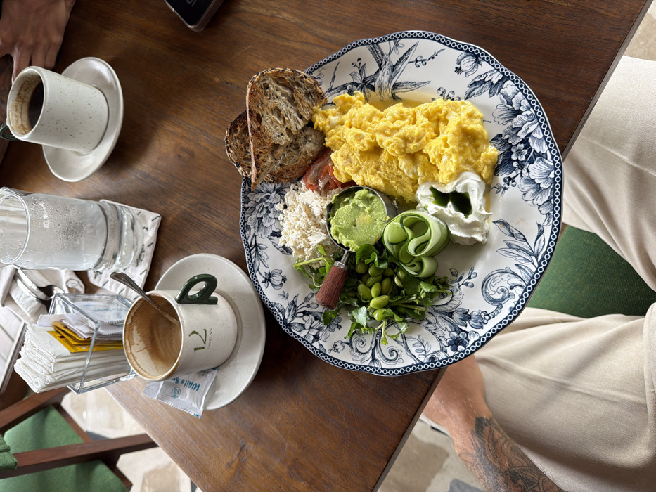 The image displays an overhead view of a breakfast or brunch setting on a dark wooden table in a cafe located in Tibubeneng, Indonesia. In the immediate foreground, the lower torso and legs of an individual wearing light-colored (beige/khaki) trousers are visible, along with a segment of a tattooed left arm resting on the table.
A prominent white plate with a blue floral pattern occupies the center, laden with various food items. These include a generous portion of bright yellow scrambled eggs, two slices of toasted dark bread, a small metal cup containing mashed avocado (guacamole), a pile of green edamame beans, several green leafy garnishes (likely watercress), rolled or folded cucumber slices, a dollop of white creamy substance (e.g., yogurt, labneh) with a green drizzle, crumbled white cheese-like substance (e.g., feta or tofu), and a few sliced red tomatoes. A small utensil with a wooden handle rests within the avocado cup.
To the upper left of the main plate, an off-white coffee cup with a green handle, marked "12 TWELVE," sits on a saucer, appearing largely empty with a spoon inside. A white sugar packet labeled "White Sugar" rests on the saucer. Adjacent to this, a clear glass container holds a stack of white paper napkins and multiple yellow sugar packets. A tall, clear glass of water, exhibiting condensation, is placed on a patterned coaster nearby.
In the upper right portion of the frame, a second off-white coffee cup, containing dark black coffee, is positioned on a saucer. A human hand and forearm are visible near this cup, suggesting the presence of a second individual. A black-cased smartphone is partially visible on the table's edge in this area. Elements of a green upholstered chair are visible in the background and partially beneath the table, reinforcing the cafe environment.