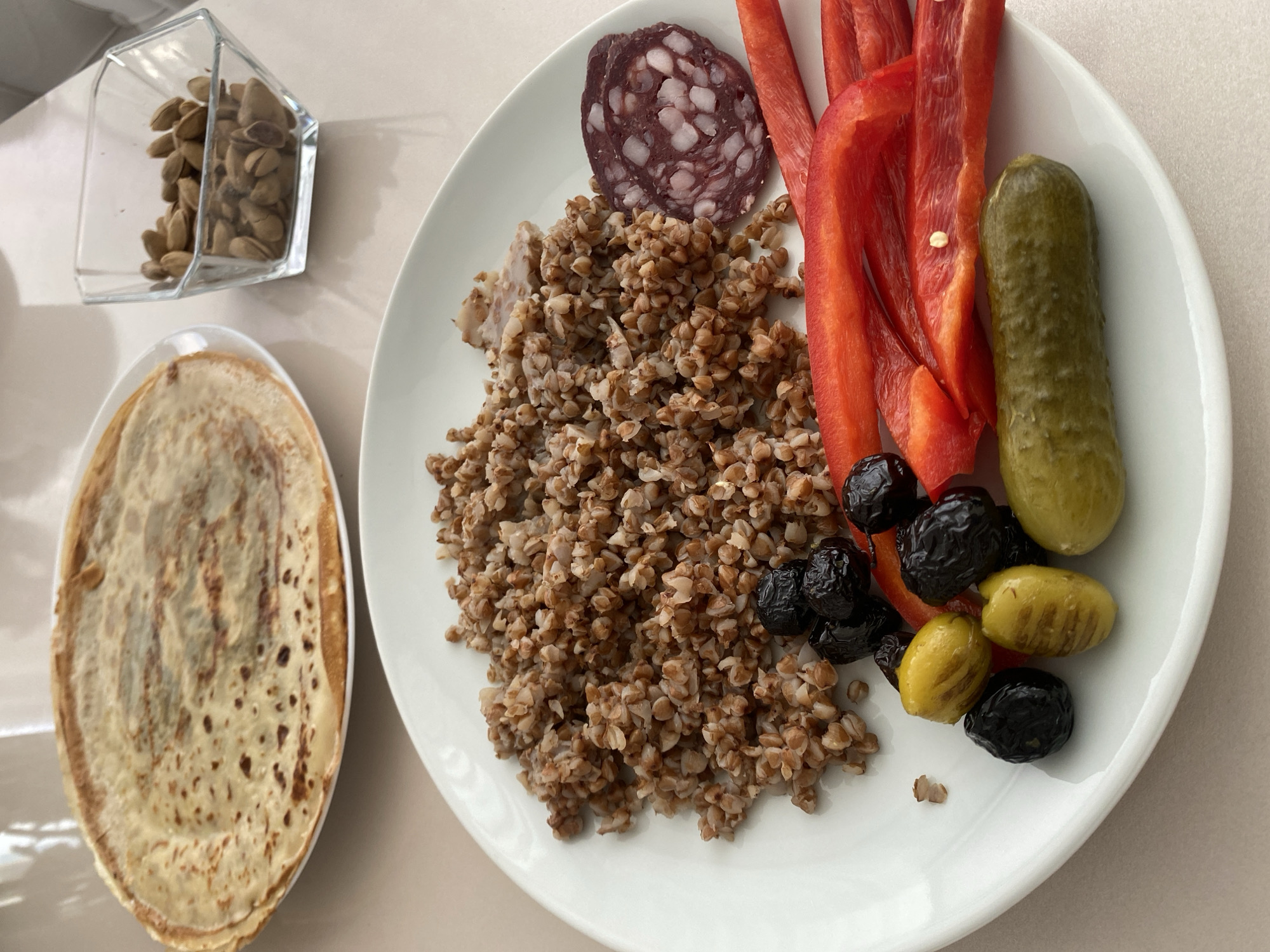 The image depicts a meal arranged on a light-colored surface. On the left, a stack of pancakes is presented in an oval dish. Adjacent to it, a small glass container holds a quantity of nuts. A white plate contains a variety of food items, including a heap of cooked buckwheat, a slice of sausage, strips of red bell pepper, a pickled cucumber, and a mix of black and green olives. The overall impression is of a simple, homemade meal. There is no information to verify the location of this image is in Alanya, Turkey.