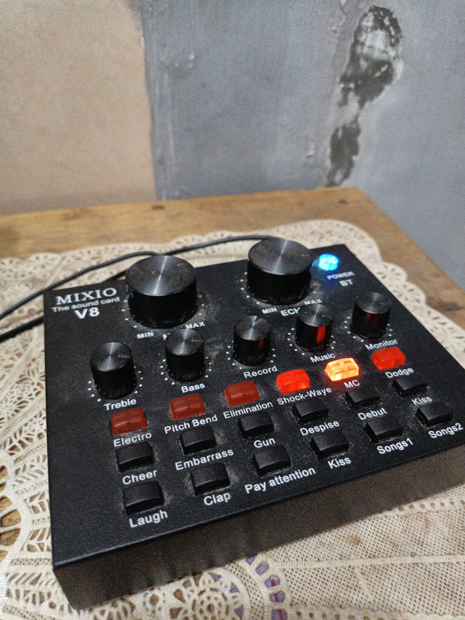 A black MIXIO V8 sound card is positioned on a wooden table covered with a white lace tablecloth. The sound card has numerous knobs and buttons, some of which are illuminated with red, orange, and blue lights. A power indicator light is blue, and several buttons like "MC," "Music," and "Monitor" are illuminated in red or orange. The background consists of a textured wall with a light brown and grey section. The scene appears to be indoors, possibly in a home or studio setting. There are no people visible in the image. The overall condition of the sound card and its surroundings suggests a domestic environment.