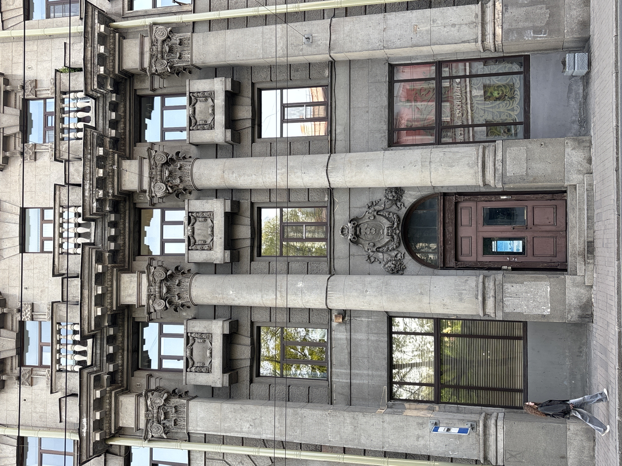The image captures a partial view of an ornate building facade in Saint Petersburg, Russia. The building features a grey stone exterior with prominent architectural details, including cylindrical columns, decorative carvings at the top of the columns, and framed rectangular elements between them. The windows vary in size and some reflect the surrounding environment. A brown door is visible, flanked by a decorative crest. In the bottom right corner, a person lies on the sidewalk, suggesting a street-level view. A street sign is also visible near the person.