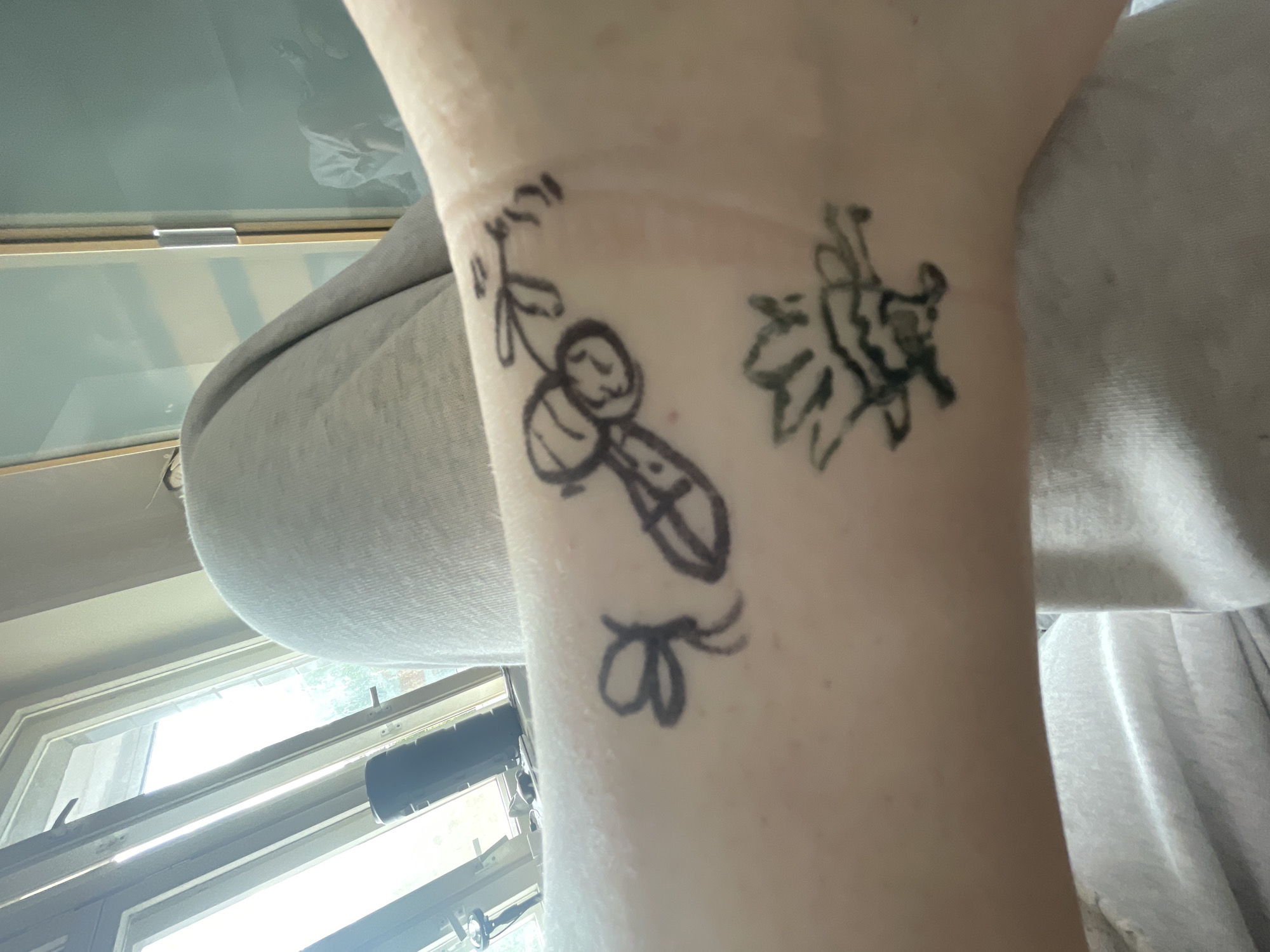 A close-up shot focuses on a person's light-toned forearm and wrist, adorned with several black ink drawings, likely temporary tattoos or marker art.
The central drawing depicts a stylized cartoon bee or insect with a round head, a striped body, and small wings, holding what appears to be a shovel or stick. Above this, a simpler drawing of a leafy sprig extends towards the wrist. To the right of the bee, a larger, more intricate floral or organic design is visible. Below the central figure, near the bottom edge of the frame, is a small, simple drawing resembling a butterfly or a ribbon.
The arm is held against a blurry background that reveals an indoor setting. A bright window is visible in the background, suggesting it is daytime and allowing natural light into the space. Hints of outdoor greenery can be vaguely discerned through the window. Portions of light-colored fabric, possibly clothing or upholstery, flank the arm on either side. The image captures a casual moment, showcasing the temporary body art. No discernible text is present in the image. While the provided context is Sinyavino, Russia, the image itself does not contain any visual cues to confirm this specific geographical location.