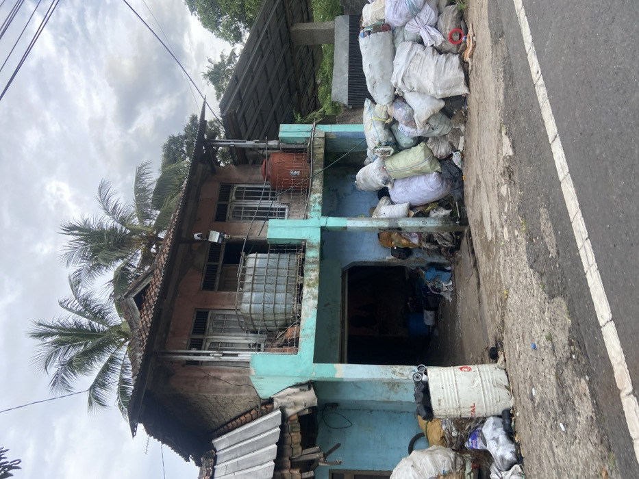 The image shows a two-story residential building with a light blue facade, a gray tiled roof, and a balcony on the second floor. The building is located on the side of a paved road in Cipatat, Indonesia, with coordinates -6.785, 107.435. The road has a white painted stripe along the edge. The building has a large open garage door on the ground floor. The garage is filled with debris and garbage bags. Outside the garage, in front of the building, there is a large pile of white garbage bags, with other trash and debris scattered around them. There is a metal water tank on the balcony. There is also a reddish-brown barrel in front of the building, with a black cylindrical container behind it. The roof of the building appears damaged and uneven, with some tiles missing. The building is surrounded by trees, and the sky is overcast.