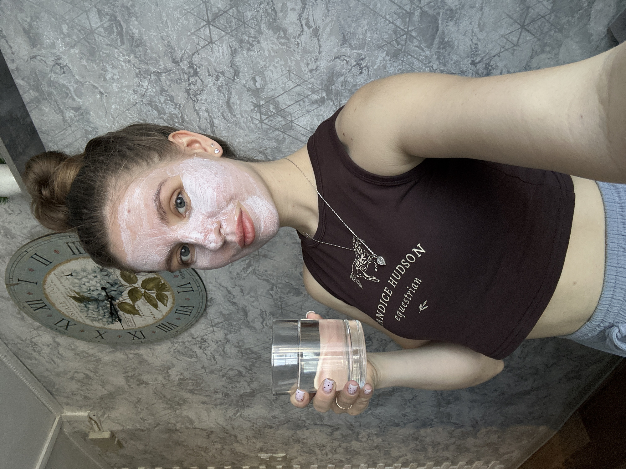 A young woman is the primary subject of this eye-level, close-up, indoor photograph. She has a white facial mask on her face. Her hair is pulled back into a bun. She is wearing a dark brown tank top with white lettering. A silver necklace is visible. In her right hand, she holds a clear glass.
In the background, a decorative clock is mounted on a textured gray wall. The overall orientation of the photo is rotated counterclockwise. The city of Klin, Russia, is not detectable.