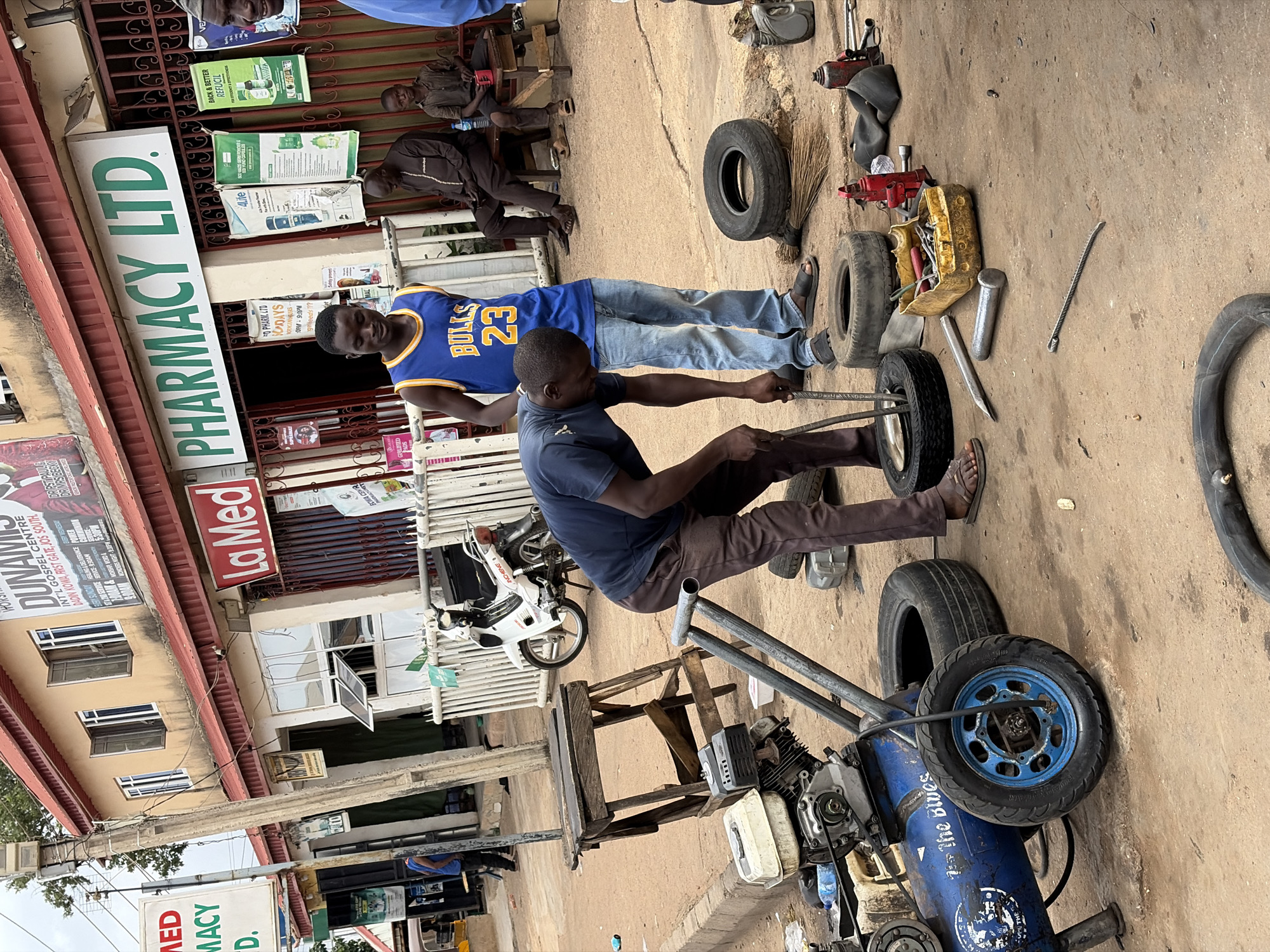 The image depicts a roadside tire repair scene, likely in Jos South, Nigeria. Two men are working on a tire, with one using a tool to pry it. A motorcycle and an air compressor are nearby. Other tires, tools, and inner tubes are on the ground. In the background, people are sitting on chairs in front of a building labeled "La Med Pharmacy LTD." A sign for "DUNAMIS INT'L GOSPEL CENTRE" is visible on the building's exterior. A man wears a "Bulls 23" jersey.