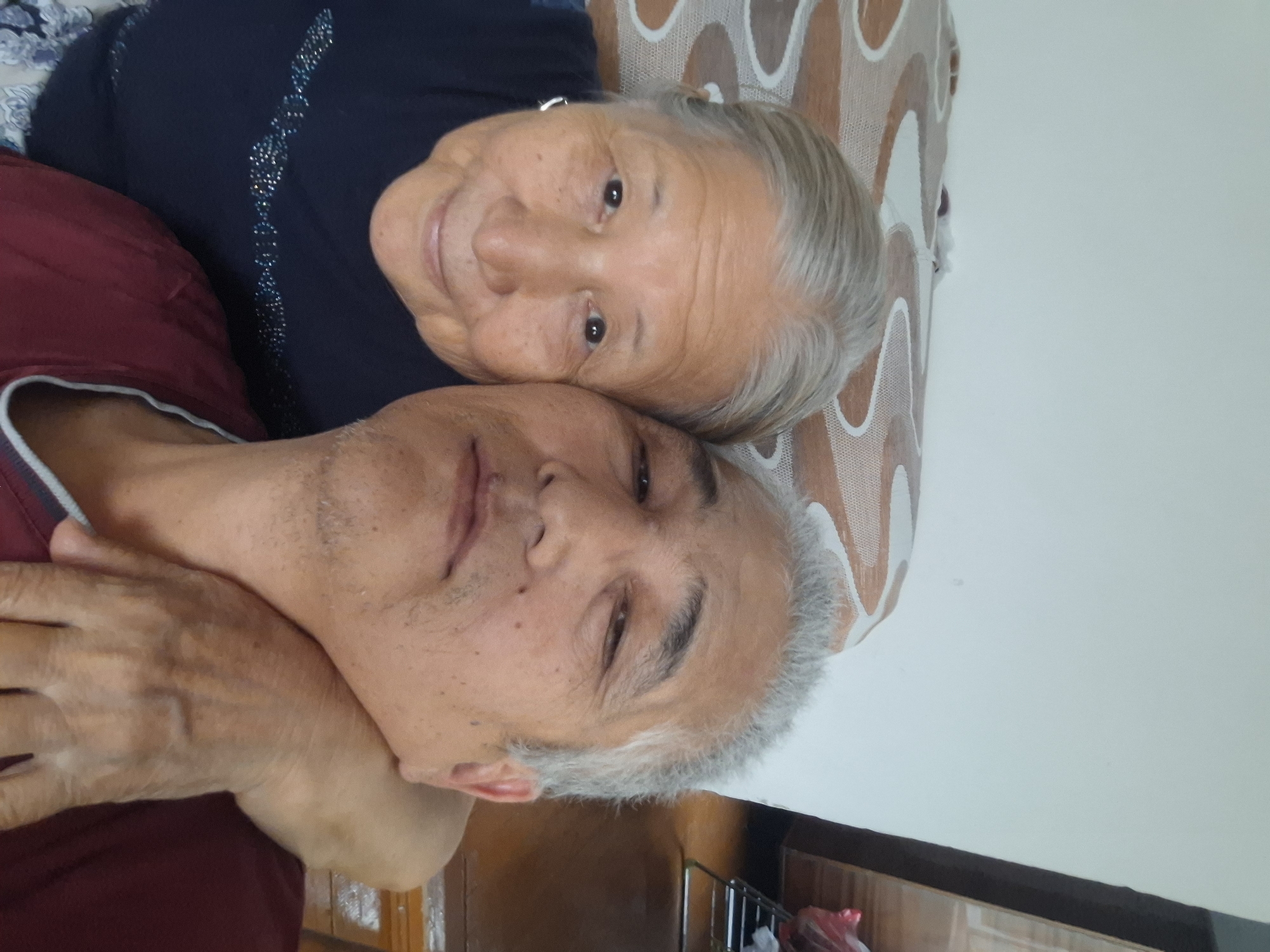 A selfie is taken in a close-up view, featuring two individuals, an older woman and a middle-aged man, likely mother and son, posing for the camera. The woman is on the left, with graying hair pulled back, a light smile, and is wearing a dark blue top adorned with a beaded pattern. The man is on the right, his arm wrapped around the woman's neck, with graying hair and a faint stubble. He is wearing a maroon shirt with a light gray neckline. They are positioned against a wall with a bed or couch in the background, covered with a patterned throw. The overall environment appears to be an indoor setting, possibly a home, in the city of Almaty, Kazakhstan. The lighting is natural and diffused, indicating it was taken during the day.