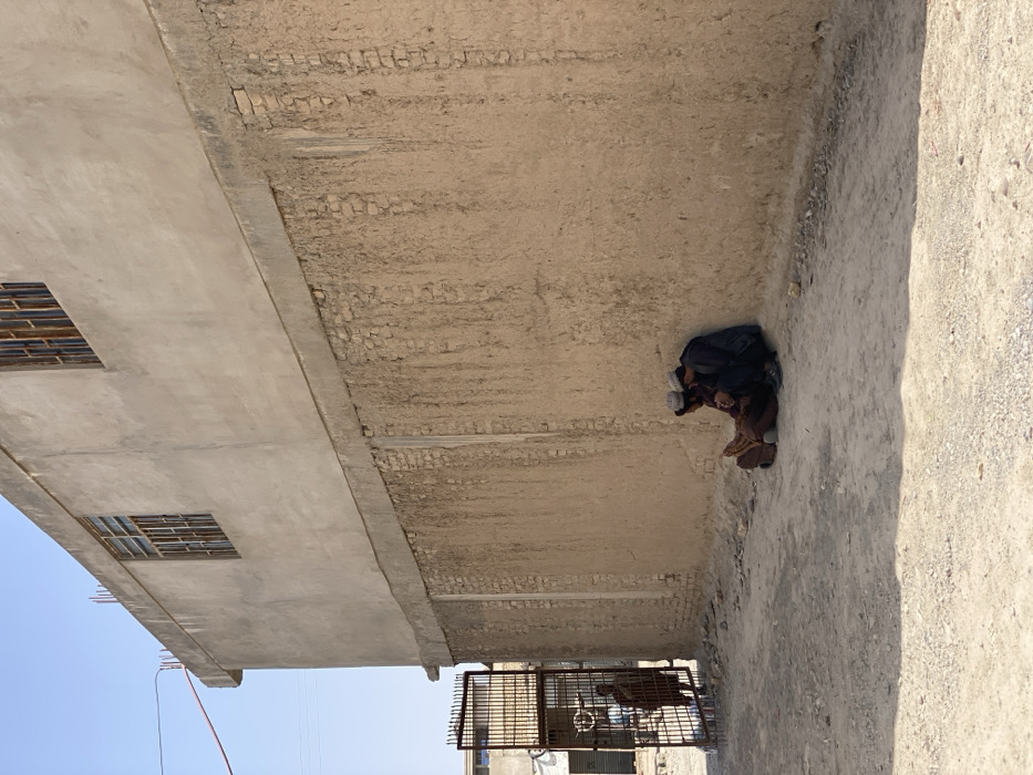 This image depicts a dusty street scene in Kandahar, Afghanistan, under clear daylight conditions. In the midground, two men are seated on the unpaved ground, leaning against the base of a tall building wall. Both men wear traditional clothing and light-colored head coverings. The man on the left is dressed in brown tones, while the man on the right wears dark blue/grey attire; they appear to be engaged in a close interaction.
The building wall consists of a lower section constructed from exposed mud brick with vertical impressions, topped by a concrete beam, and an upper section finished with light-colored plaster. Two windows with metal bars and frames are visible on the upper plastered section. To the left of the wall, a large, dark metal gate stands partially open, revealing a third individual in a dark reddish-brown garment standing just beyond it, facing away from the primary view. Beyond the gate, other structures and what appears to be a marketplace or street are visible under a clear, pale blue sky. A red cable diagonally traverses the upper left, with other utility wires visible in the distance. The ground is consistently unpaved and scattered with small debris.