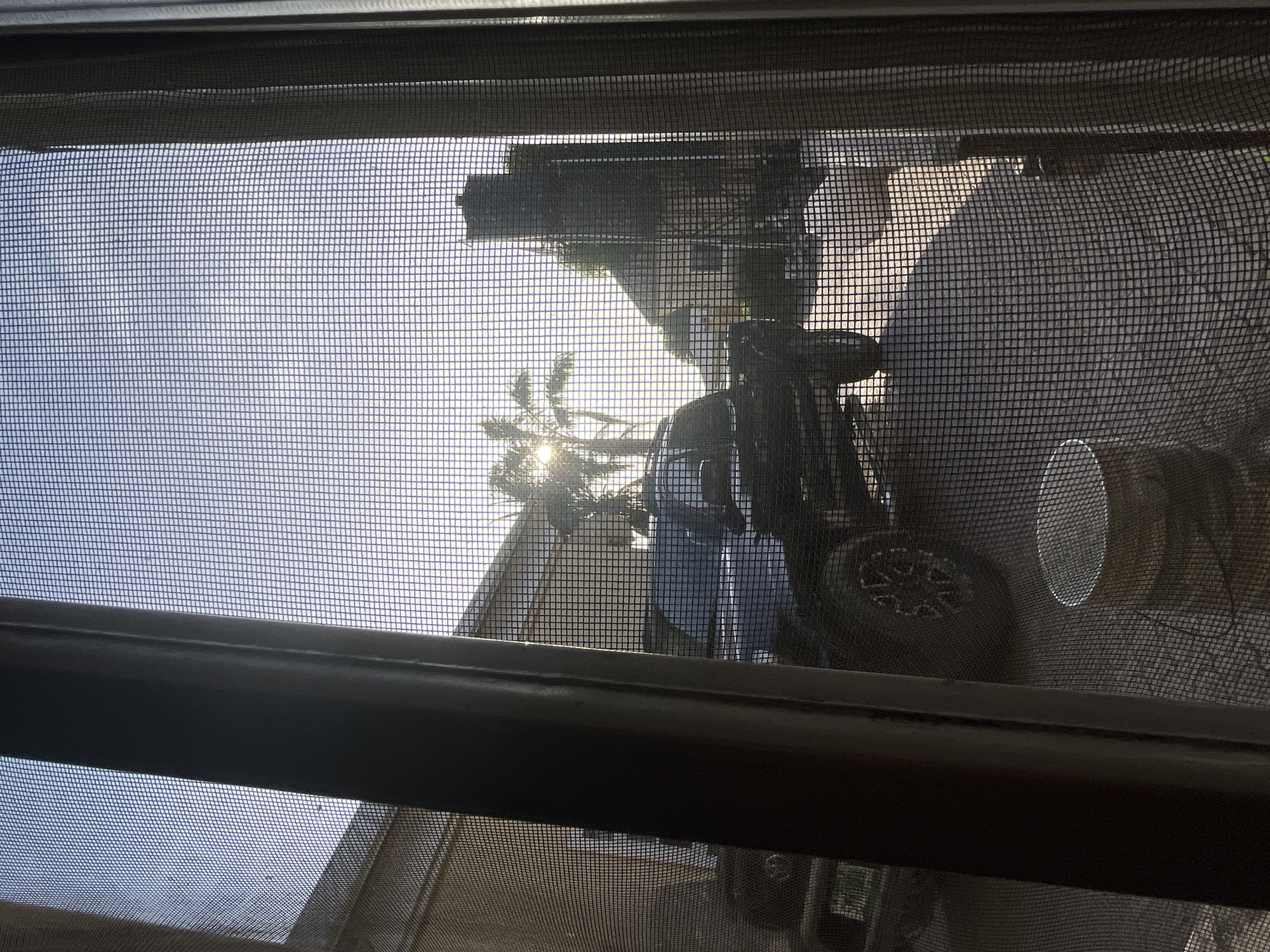 The image presents an outdoor scene viewed through a window screen, which dominates the foreground with its dark, grid-like pattern. Sections of a dark window frame are visible at the top, bottom, and as a horizontal divider bar across the middle.
The bright outdoor setting suggests a clear, sunny day, with intense sunlight. The sun itself is visible as a strong, starburst-like light source in the upper left portion of the image, partially obscured by the dark silhouette of a palm tree whose fronds are backlit.
In the mid-ground, a large, dark object resembling a recreational vehicle or utility equipment is prominent. A distinct wheel with a multi-spoke design is visible, along with parts of the vehicle's body and what appear to be handlebars or controls. Other dark, indistinct shapes in the background suggest additional structures, vehicles, or equipment. A light-colored, circular, possibly reflective object is also discernible towards the right side.
No specific activity or event is taking place, and no people are visible in the frame. There is no discernible text. The bright sun and presence of a palm tree are consistent with a warm climate, aligning with the provided location context of Abuja, Nigeria.