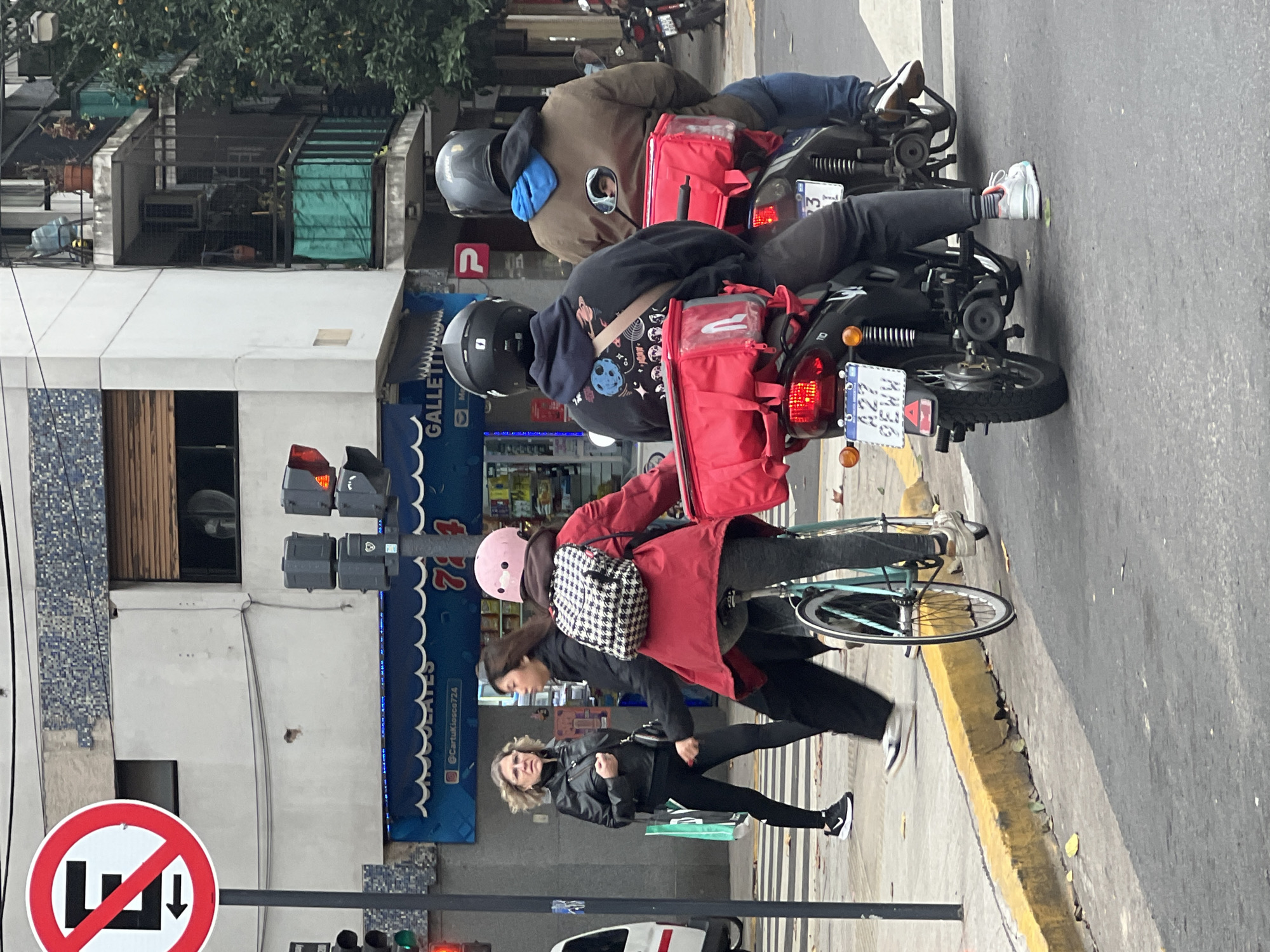 A street scene in Buenos Aires, Argentina, shows two delivery motorcyclists stopped in the street next to a traffic light. Both riders wear helmets, and each motorcycle has a red delivery container attached. The vehicle license plates are visible. A woman rides a bicycle past them, also carrying a delivery container. A pedestrian walks on the sidewalk parallel to the cyclist. A "No Parking" sign is visible on a post near the traffic light. Buildings with storefronts line the street.
