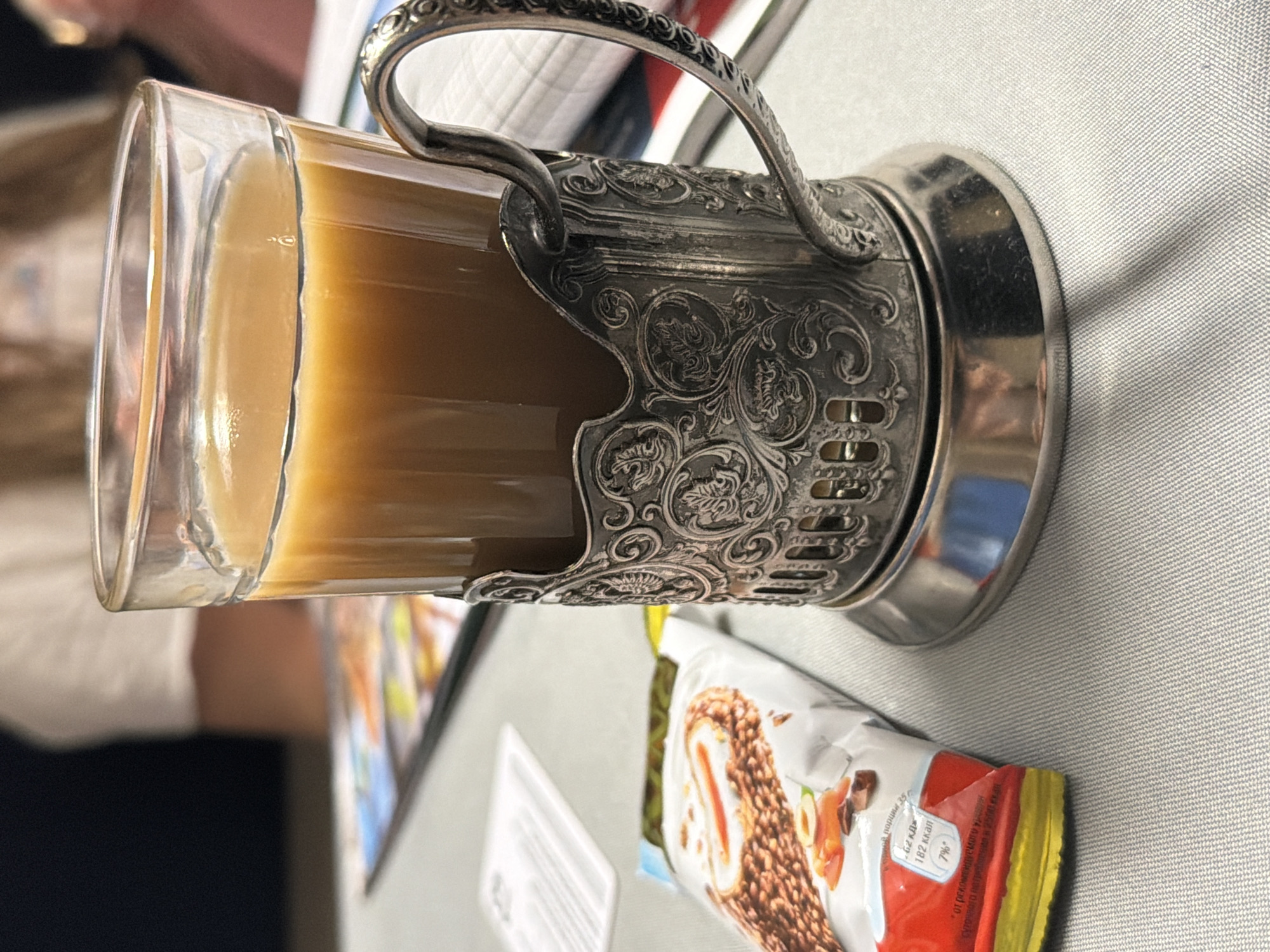 An indoor scene in Kostino, Russia, displays a glass cup containing a light brown beverage, likely coffee or tea with milk, seated within an ornate, silver-colored metal podstakannik featuring embossed floral and leaf motifs. To the left, a partially open Kinder-brand snack wrapper, predominantly white and red, displays an image of a chocolate bar with nuts and caramel, along with Russian text. All items rest on a light grey, subtly textured fabric surface. The background is out of focus, revealing blurred figures of individuals and indistinct papers or booklets on an adjacent surface. No direct human action is depicted within the frame.