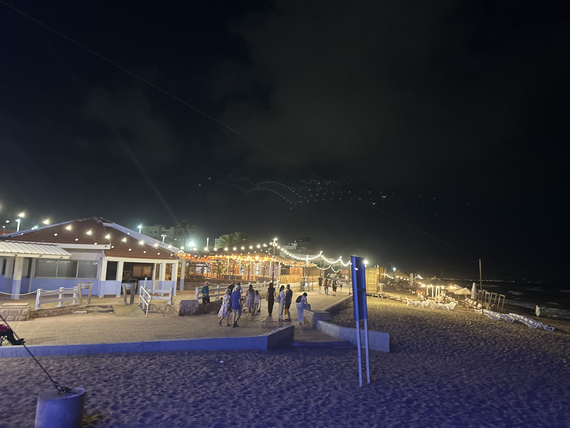 This image depicts a nighttime scene on a sandy beach in Nahariyya, Israel. A boardwalk lined with white lounge chairs and umbrellas runs along the edge of the sand. Further inland, a building with a red-tiled roof is illuminated by strings of warm lights, and several people are seen walking on the boardwalk in front of it. The sky above is dark and cloudy, with a thin line of lights visible on the horizon. The beach itself is sparsely populated, with a few individuals scattered across the sand.