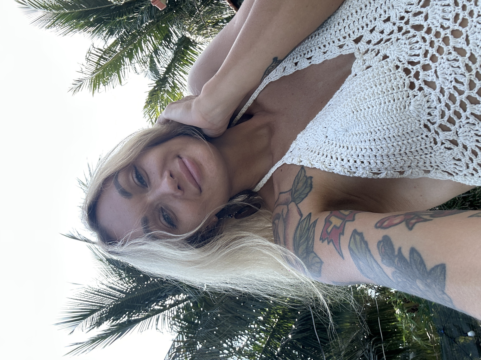 A woman with blond hair is centered in a low-angle selfie with palm trees visible in the background, suggesting an outdoor location. She is wearing a white, crochet-style top. The location appears to be Mũi Né, Vietnam, based on the vegetation and overall atmosphere. The woman has tattoos on her arms. Her pose and facial expression suggest a casual and relaxed setting.