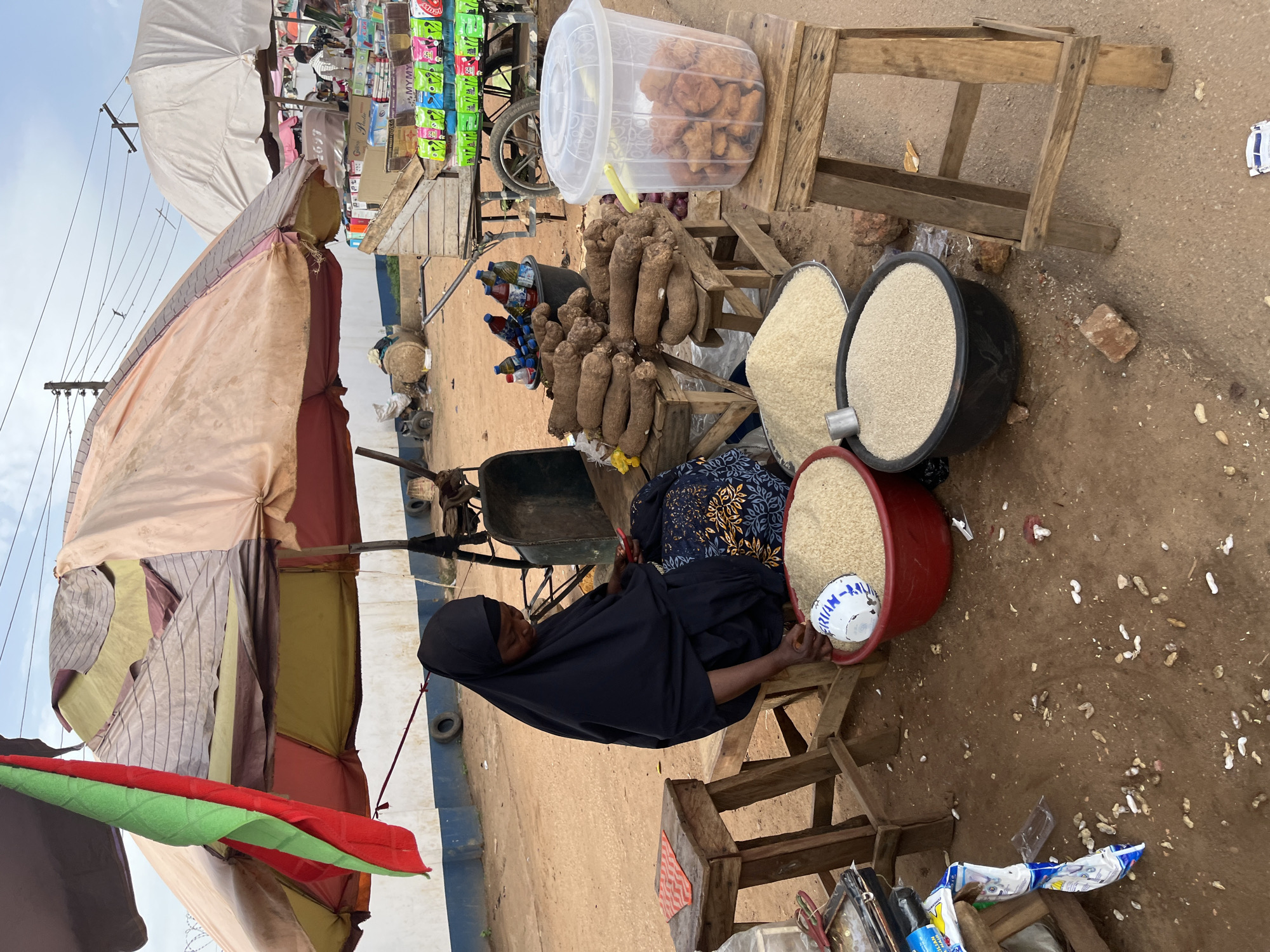 The image depicts an outdoor market scene in Rafin Par, Nigeria. A woman wearing a black hijab and traditional attire is seated on a wooden stool, engaged with her phone. She is positioned in front of a stall covered by a large, multi-colored umbrella. Various goods are displayed, including large bowls of rice, yams, and a clear plastic container filled with fried snacks. In the background, a cart is laden with packaged goods, and other stalls and market activities are visible. The ground is dusty, and the overall environment suggests a typical bustling African marketplace.