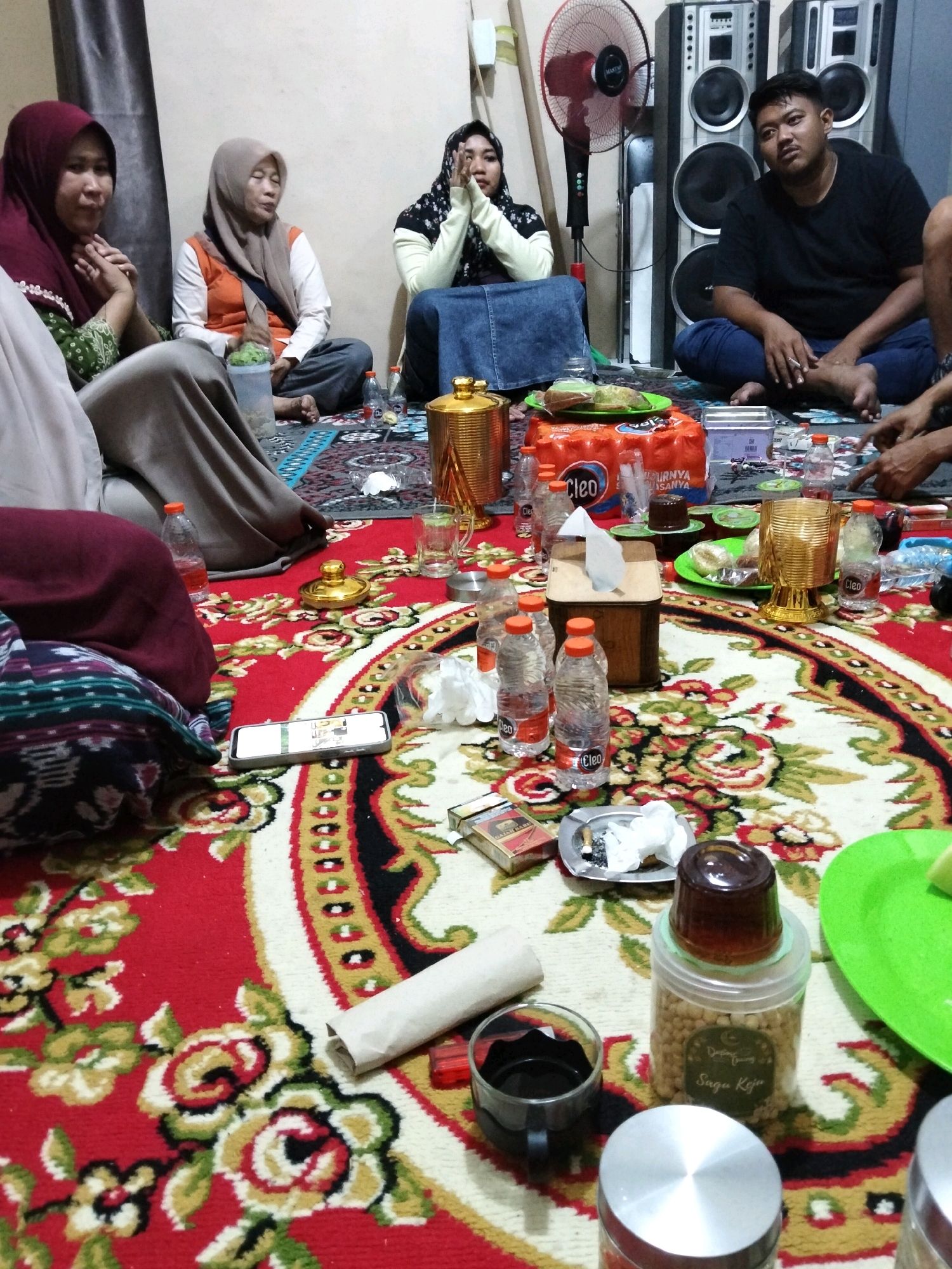 In a room with a carpeted floor, a group of people are gathered. The room appears to be a living space, with a fan and stereo speakers visible in the background. In the foreground, a red carpet with a floral pattern is laid out. Several individuals are seated on the carpet, some wearing hijabs. A table is set up with various items, including bottles of water, snacks, a tissue box, and smoking accessories. The overall scene suggests a casual gathering or social event. The location is identified as Puskesmas Keboan, Indonesia.