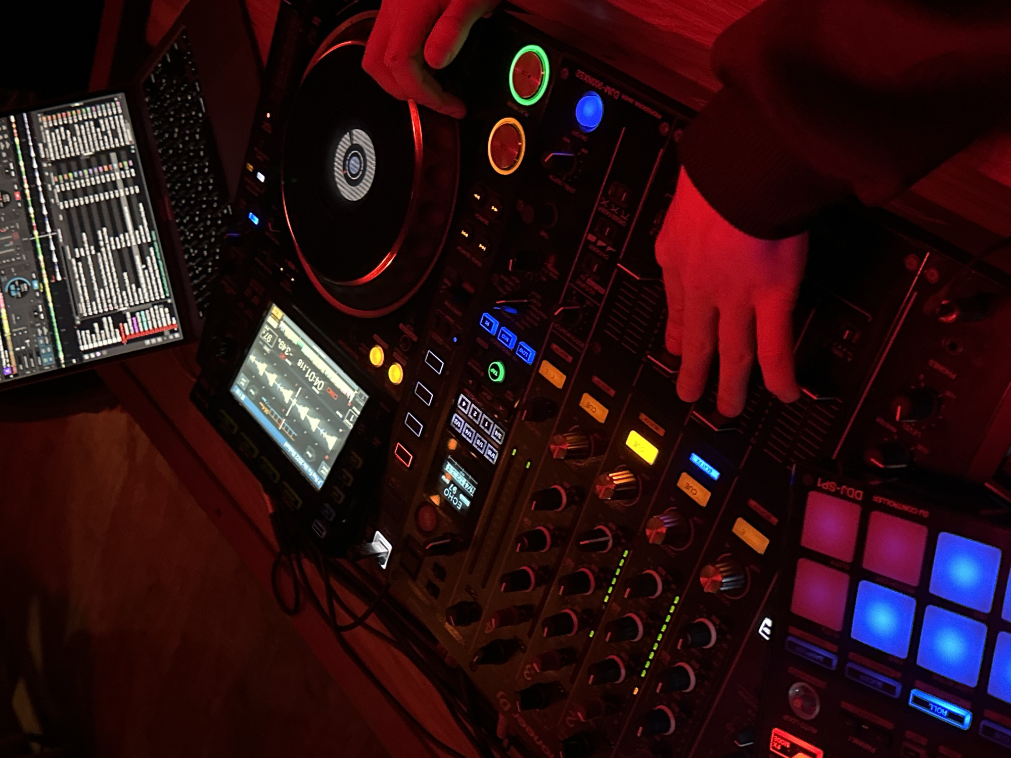 A DJ console is seen in a dimly lit, possibly nightclub or studio, environment. The console has numerous buttons, knobs, and a screen displaying waveforms and track information. A laptop with DJ software running is to the left of the console. Two hands are visible, one on the deck and the other on a mixer fader. The brand "Pioneer DJM-900NXS2" is visible on the console. A Pioneer DDJ-SP1 controller is attached to the console on the right. The location in the city of Moscow, Russia, cannot be determined from the visual data provided.
