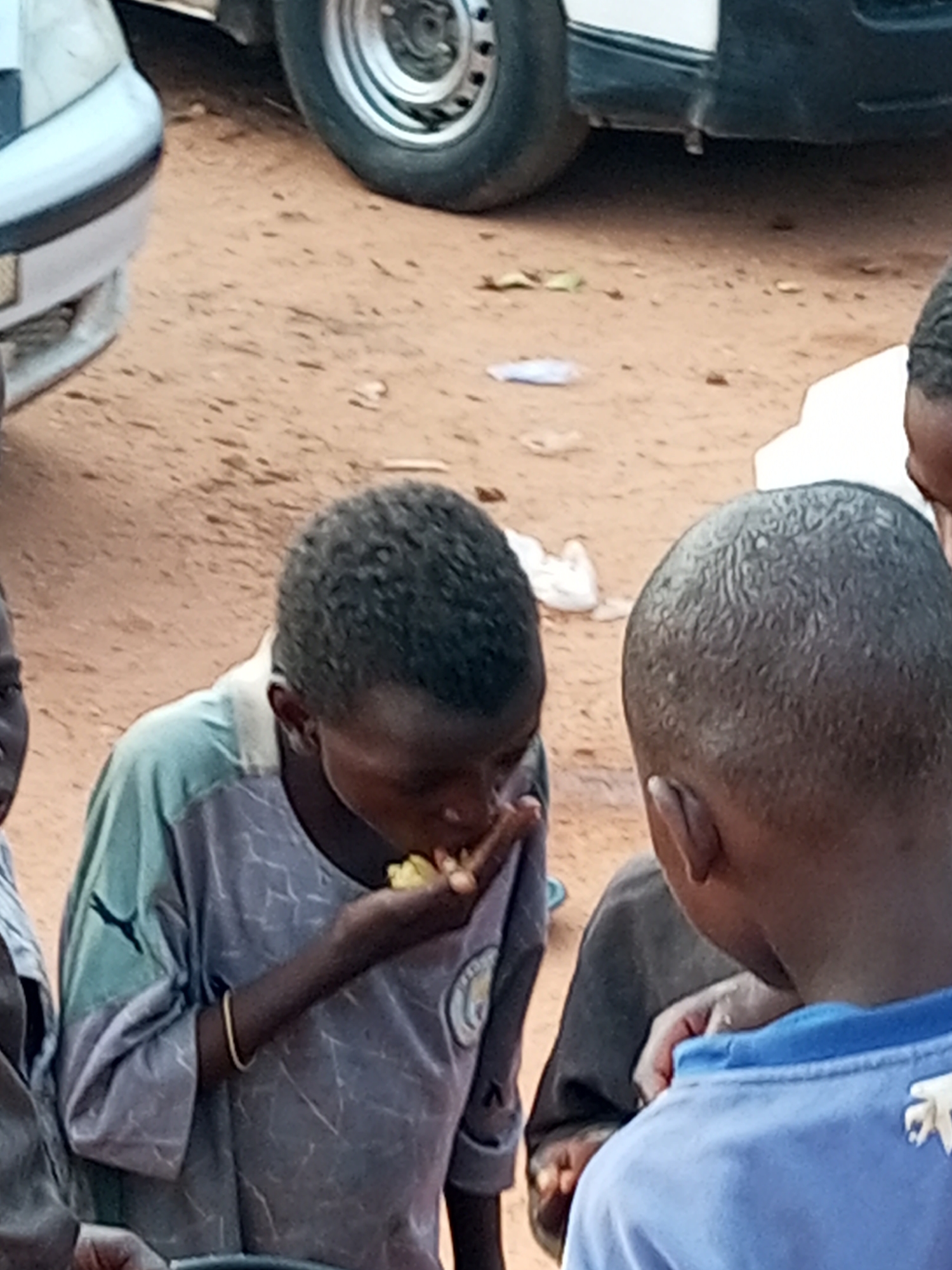 This media file captures a candid moment of children gathered around on a sandy surface, likely in Gombe, Nigeria. Three young individuals are prominently visible, with their faces partially obscured. One child in the foreground, wearing a light blue t-shirt, is seen from behind, their attention directed downwards. To their left, another child, with dark curly hair, is actively eating something yellow, possibly corn or fruit, with their right hand. This child is wearing a grey t-shirt with a Puma logo on the left sleeve and a circular emblem on the right side. A third child is partially visible to the far left.
In the background, a portion of a white vehicle is visible, with a silver wheel and tire. The ground is composed of reddish-brown sand with scattered dried leaves. A small piece of white paper or plastic can be seen on the ground further in the background.
The scene appears to be set outdoors during daylight, though the specific time of day and weather conditions are not discernible. The overall mood seems casual, with the children focused on their activity. The image is somewhat grainy, suggesting it might have been taken with a mobile phone or in low light conditions. No discernible text is present in the visible portions of the image, other than the Puma logo on one child's shirt.