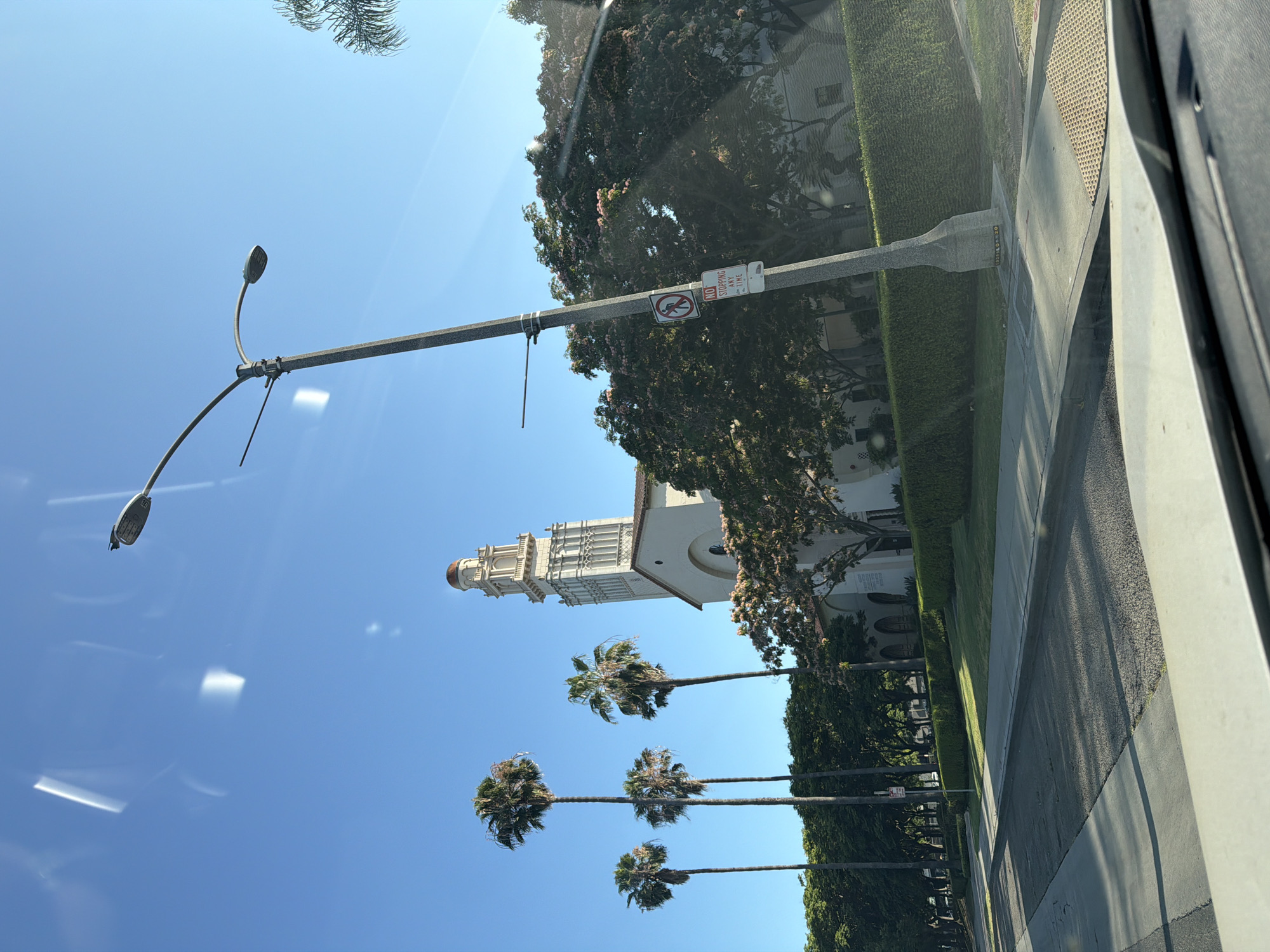 An exterior, eye-level, wide shot shows a street scene in front of a large, ornate building with a tall tower. The building is light-colored with arched windows and intricate architectural details on the tower. Several tall palm trees line the sidewalk in front of the building, with lush green bushes and trees also visible. A street light with a modern design stands in the foreground to the right. Two traffic signs are attached to the streetlight pole; one is a red and white circular sign with a pedestrian icon, and the other is a white rectangular sign that reads "NO STOPPING ANY TIME." The sky is clear and bright blue, and the sun casts shadows on the asphalt street. The view appears to be taken from a vehicle.