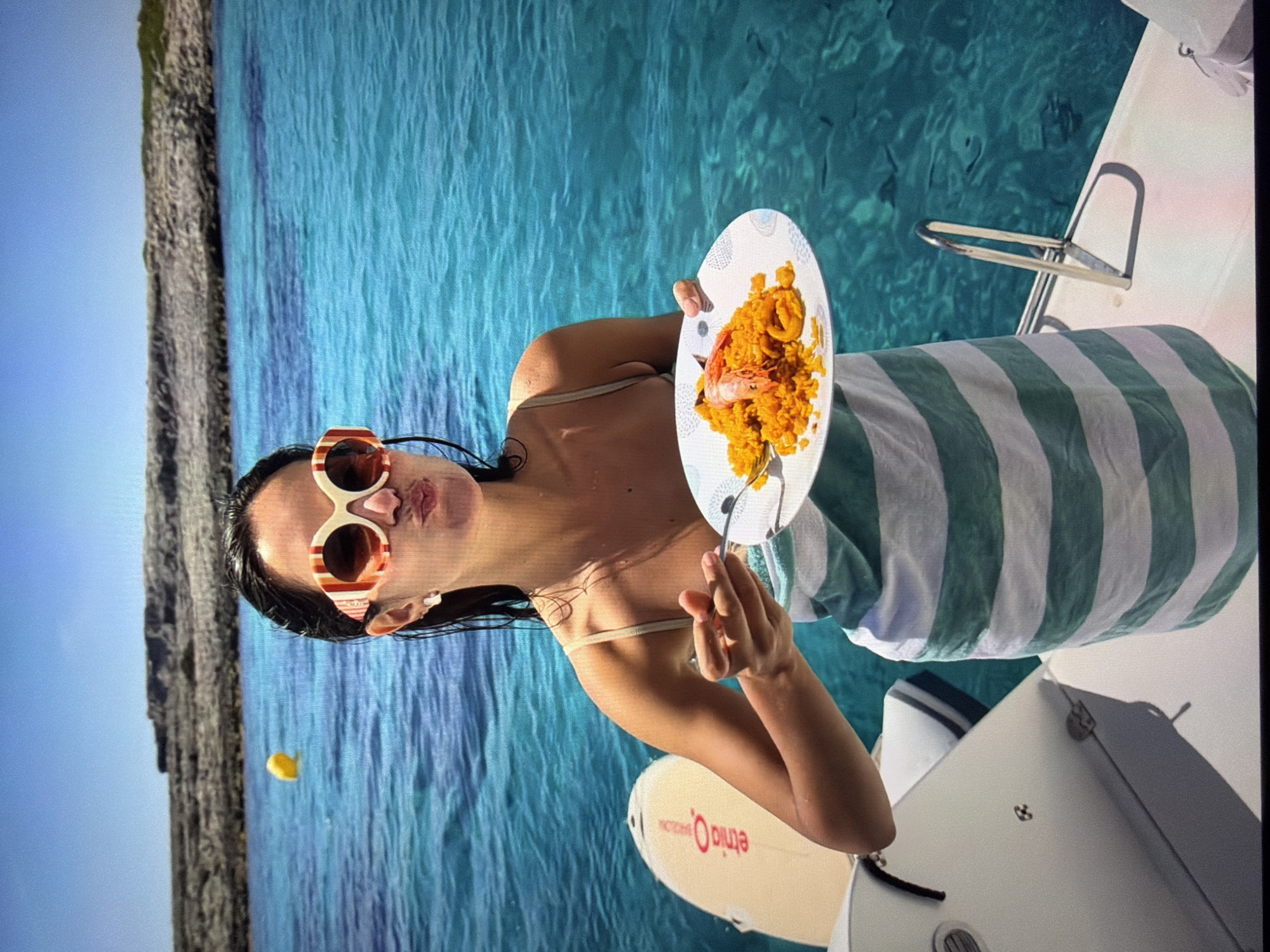 The image is oriented sideways, rotated 90 degrees counter-clockwise. It depicts a woman on a boat enjoying a meal amidst clear, calm turquoise waters. The woman, with dark hair and distinctive red and white striped sunglasses, is holding a white plate filled with an orange-yellow dish, likely paella, garnished with a large shrimp, and a fork. A green and white striped towel is draped around her, suggesting a relaxed setting. She is looking towards the camera with a slightly pouting, playful expression.
In the background, a rugged, rocky coastline meets a bright blue sky, enhancing the serene marine environment. The bright daylight indicates it is midday. A small yellow object floats in the distant water. Partially visible near the boat is a white paddleboard or surfboard with reversed red text that reads 'ełmo O.' and 'eimio' (when viewed in the current orientation). The scene, characteristic of a warm and sunny climate, is set with the provided context of Ros Na Rí, Ireland.