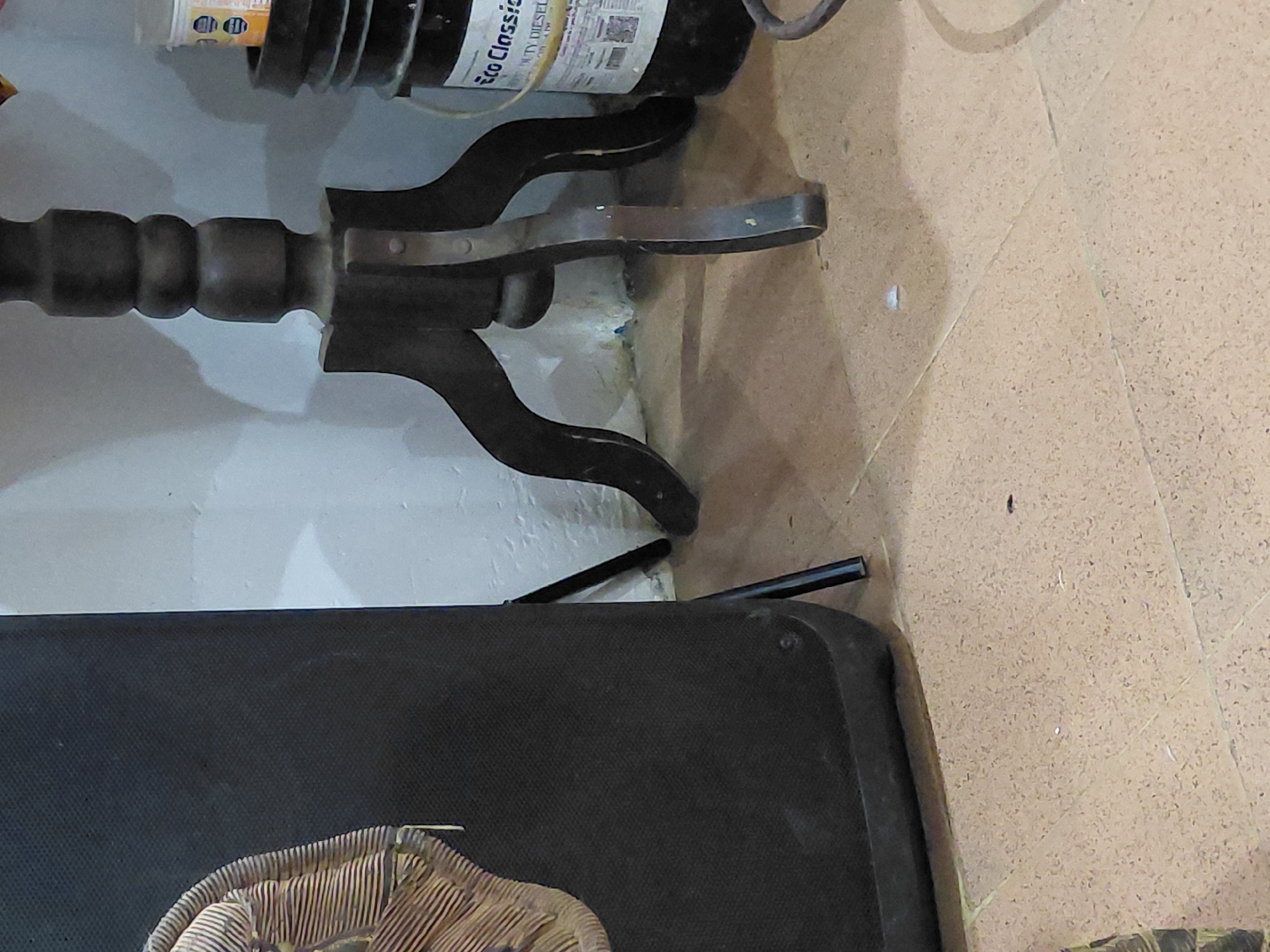 This indoor image captures a static scene in what appears to be a room corner in Santa Ana, Venezuela. A prominent dark-colored, possibly wooden, furniture leg or stand with carved, rounded segments and decorative supports occupies the central and upper-left portion of the frame. It rests against a plain white wall that shows some marks or discoloration near the base.
Above the furniture leg, a dark bottle or container is visible with a white label. Partially legible text on the label includes "Eco Classic" and "DUTY DIESEL," along with other smaller, unreadable text.
The floor is covered with light-brown or beige tiles, displaying faint grout lines and a speckled texture. In the bottom-left corner, a large, dark, rectangular object, possibly a suitcase or a fabric-covered box, is present. A portion of what appears to be a woven or braided item, possibly part of a decorative object or a fan, rests on top of this dark object. Two thin, black cylindrical rods are visible on the tiled floor, positioned between the furniture leg and the large dark item.
There is no discernible human activity or event taking place. The lighting suggests an indoor setting, likely illuminated by artificial light. No specific time of day or weather conditions can be determined from the image.