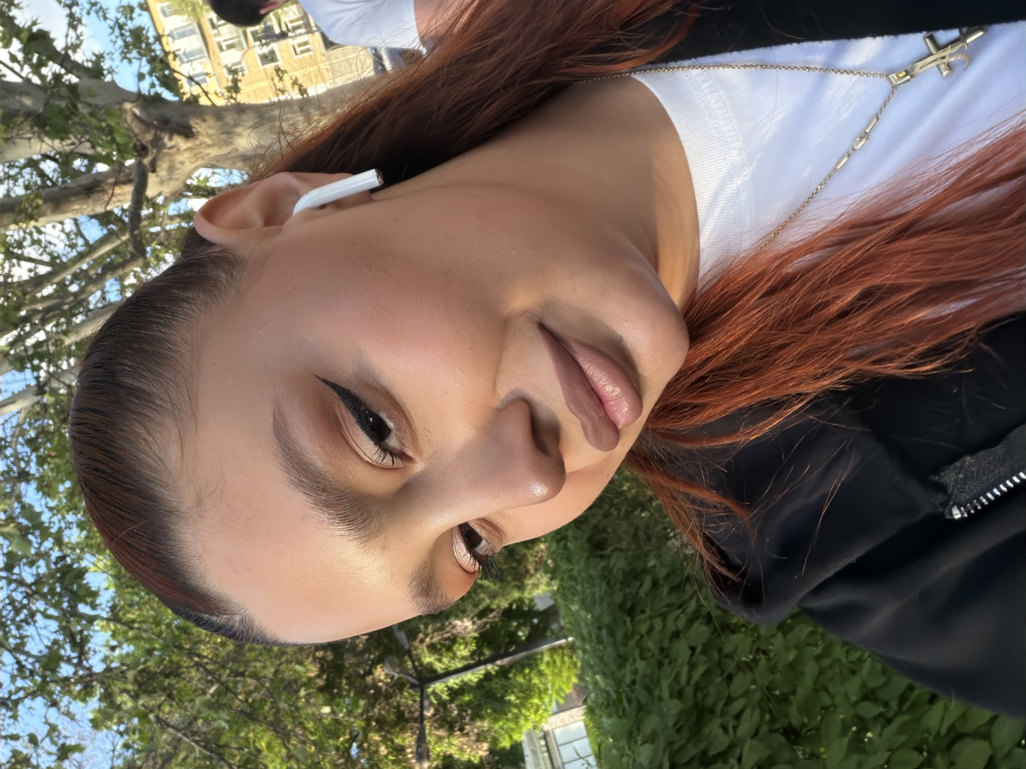 A medium close-up, eye-level, rotated view captures a young woman outdoors in a park-like setting. She has reddish-brown hair, fair skin, and wears an in-ear headphone in her left ear. Her makeup is visible with defined eyeliner. She wears a white top with a gold-colored necklace and a black jacket.
In the background, green foliage and a street lamp indicate an outdoor location. A tree and a building with windows are partially visible in the background. There are no clear indications that the location is Yerevan, Armenia.