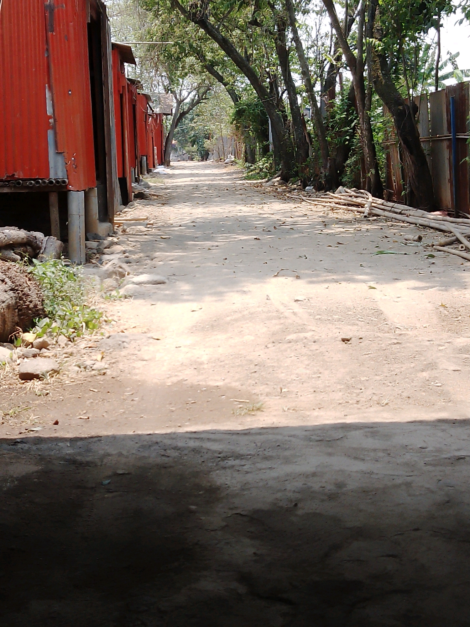 The image shows a dusty, unpaved road lined with makeshift structures on one side and trees and a fence on the other. The structures on the left appear to be made of corrugated metal, painted red, and elevated on stilts. Scattered debris and vegetation are visible near the road. The road itself is dry and appears to be a rural or semi-urban area. The lighting suggests it is daytime, with shadows cast by the trees and structures. The scene is likely in the city of Rembun, Indonesia, based on the provided context. There are no people visible in the image, and the overall impression is one of a simple, possibly impoverished, living environment.