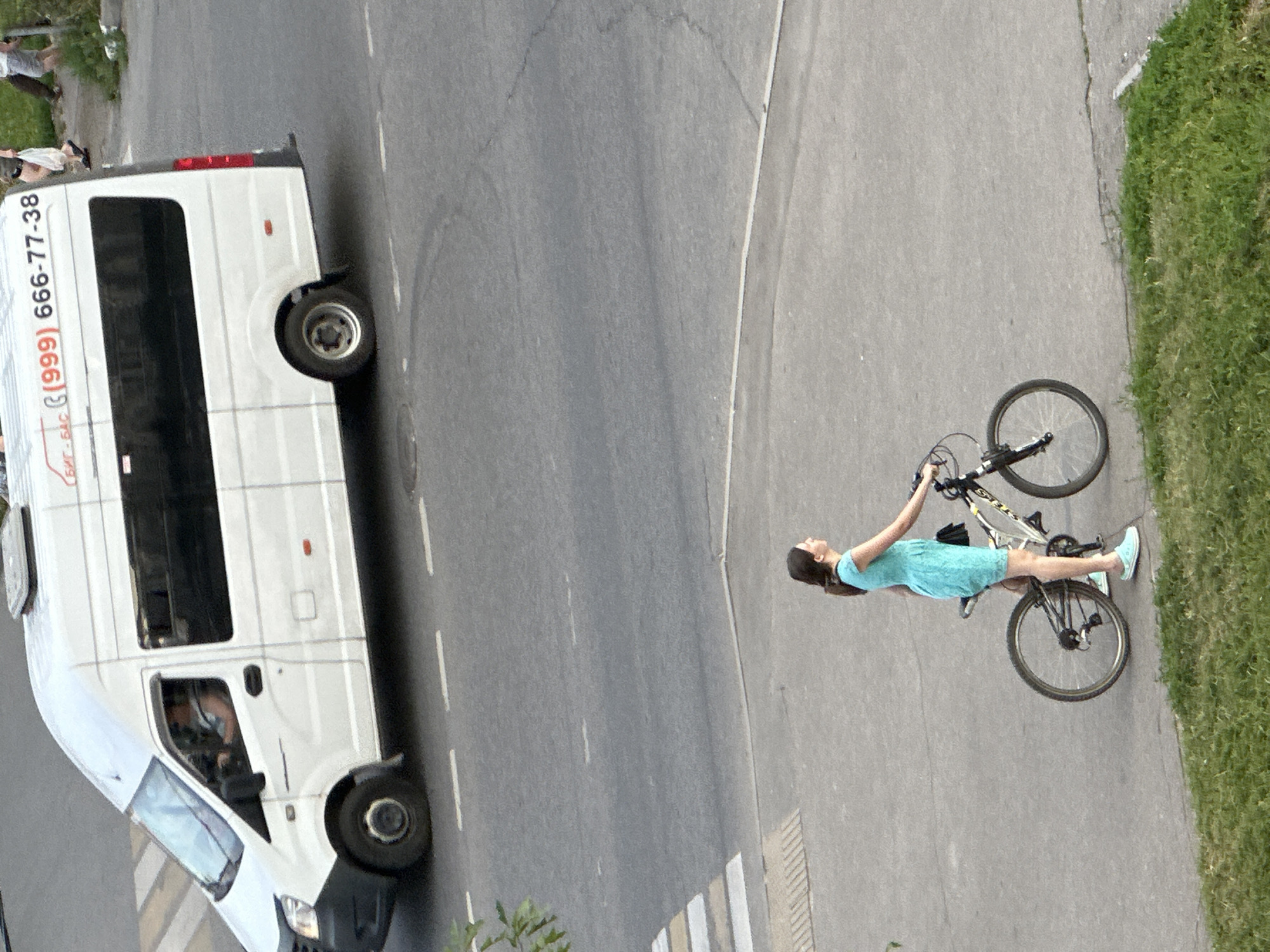 In Podolsk, Russia, a white passenger van with tinted windows and a phone number visible on its side, "(999) 666-77-38", is parked on an asphalt road. Nearby, a young woman wearing a turquoise dress and white sneakers stands next to her bicycle on a paved walkway. The bicycle is black with white accents and has a black handlebar bag. The scene is viewed from a high angle, showing the road, walkway, and a patch of green grass in the foreground.