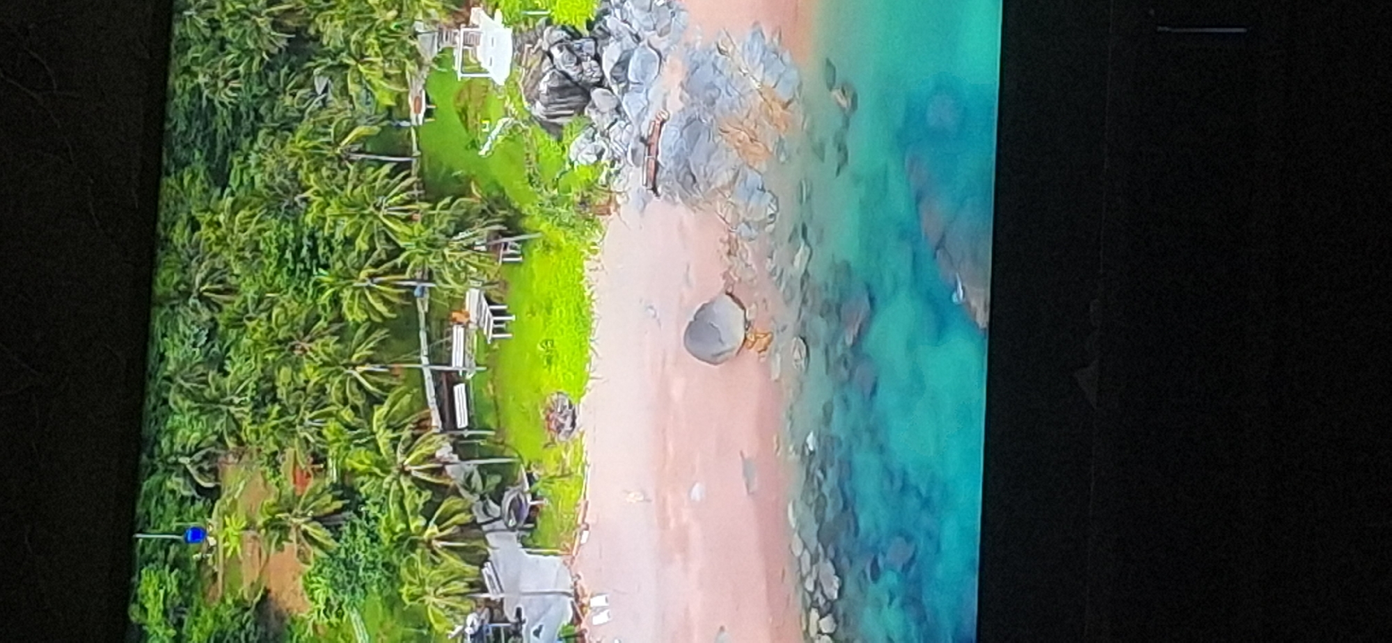 The image shows an aerial view of a tropical beach with lush green vegetation, including palm trees, and a body of water with clear turquoise waters. There are several structures on the beach, including what appears to be seating areas with tables and benches, and some covered pavilions. A large gray rock sits on the sandy shore near the water. The scene depicts a resort or a recreational area in a tropical climate. The question about the location in Bila Tserkva, Ukraine is irrelevant as the image clearly depicts a tropical environment, not the landscape of Bila Tserkva, Ukraine.